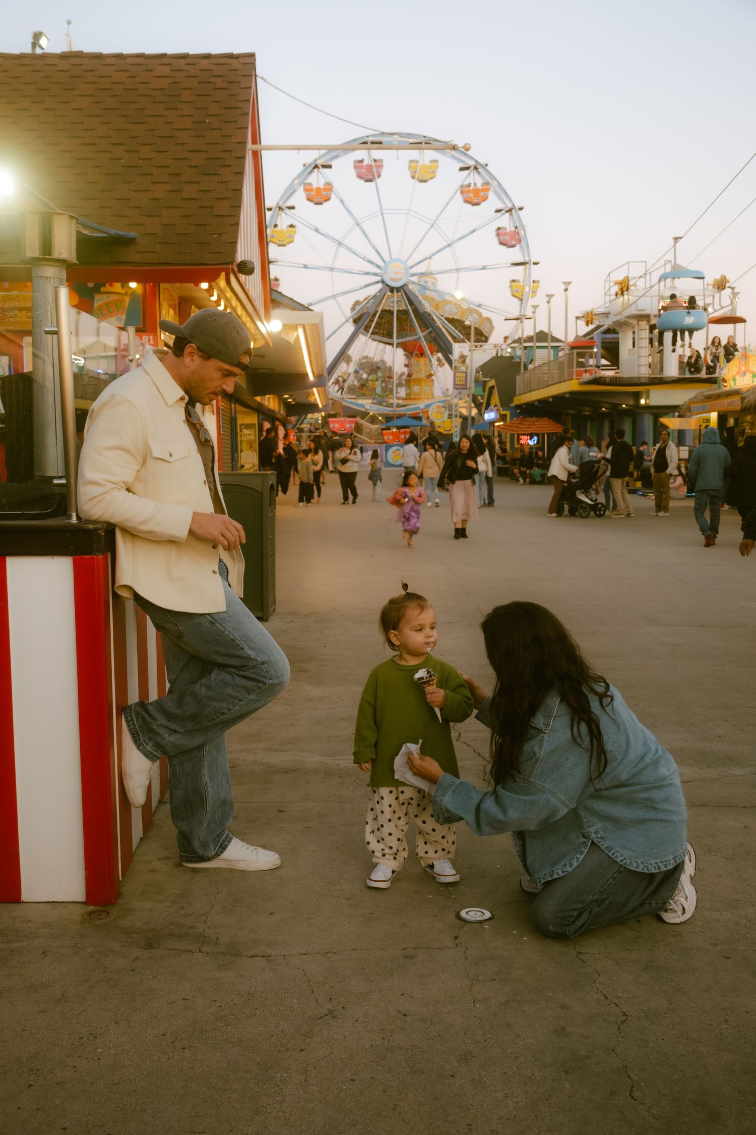 Family enjoying an amusement park at dusk, with a Ferris wheel in the background and a toddler being attended to.