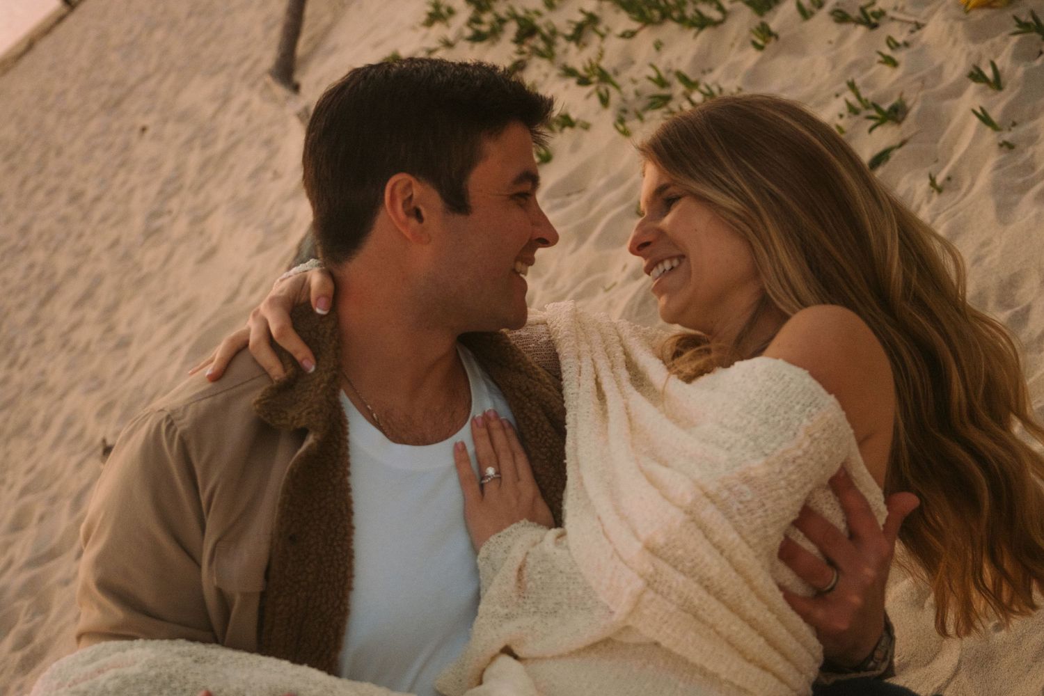 Couple smiling at each other on a sandy beach, woman wearing engagement ring, romantic golden hour setting.