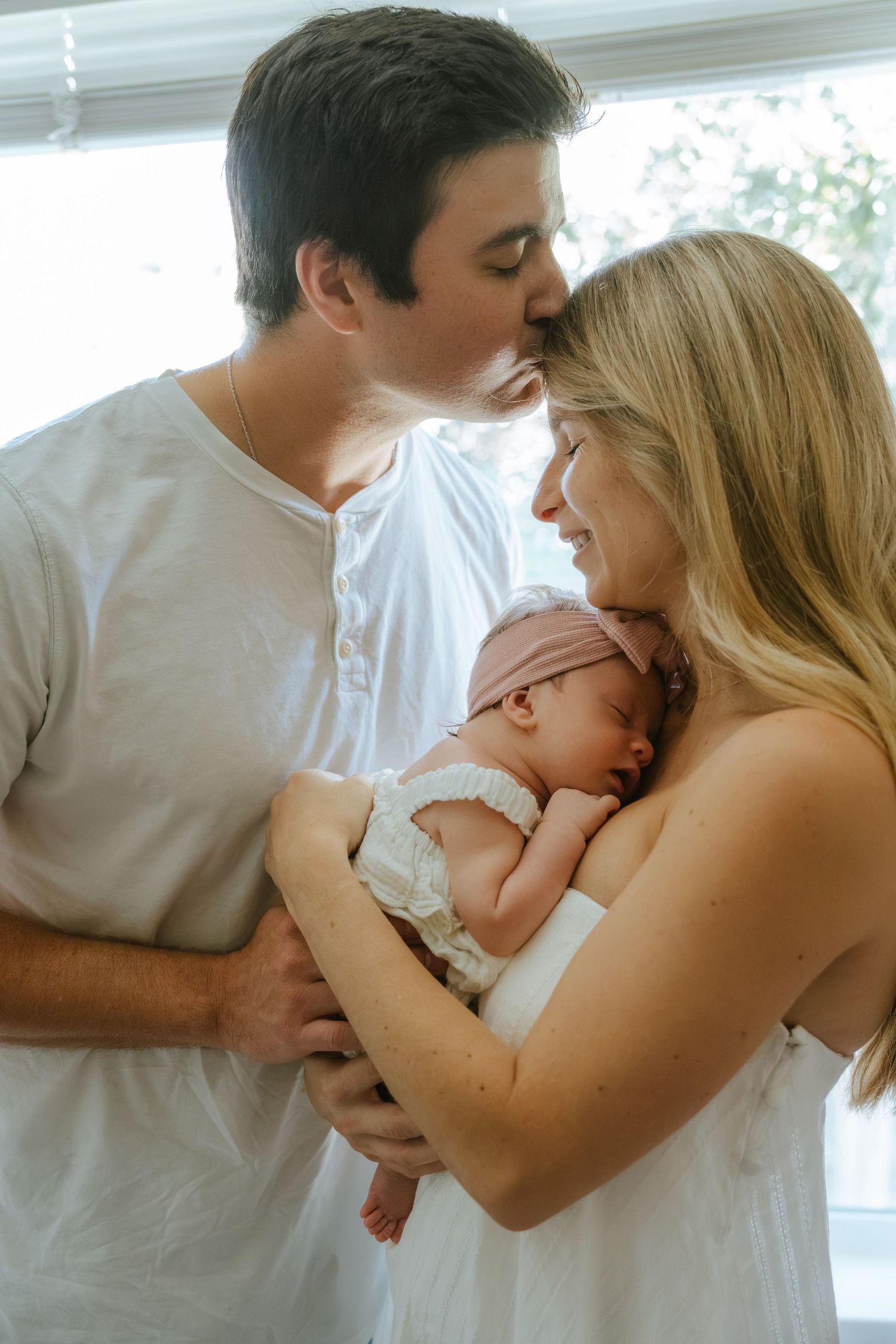 New parents share a tender kiss over their sleeping newborn baby, wrapped in white lace and a pink bow headband.