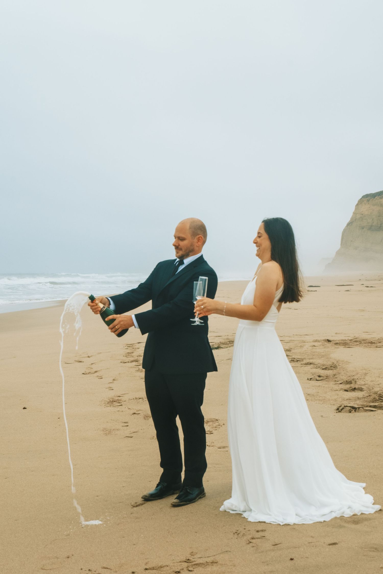 Bride and groom popping champagne on a sandy beach, celebrating their wedding day by the ocean.