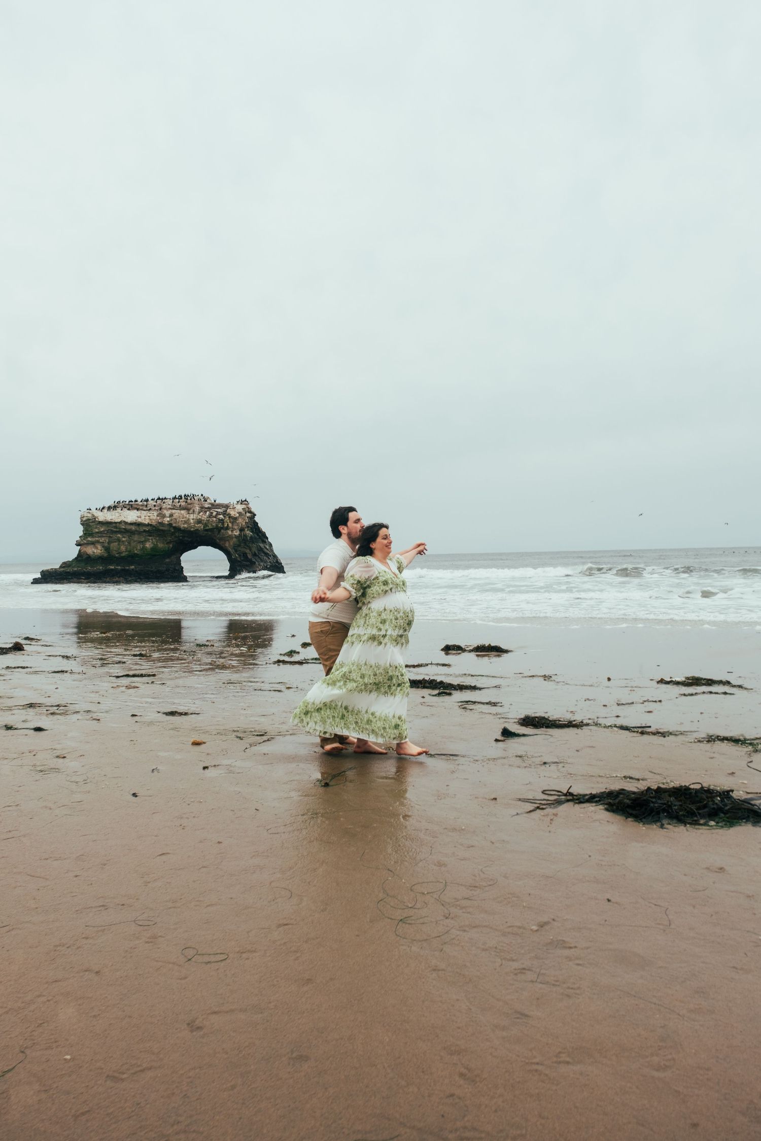 Couple dancing on a sandy beach with natural rock formations and ocean waves in the background.