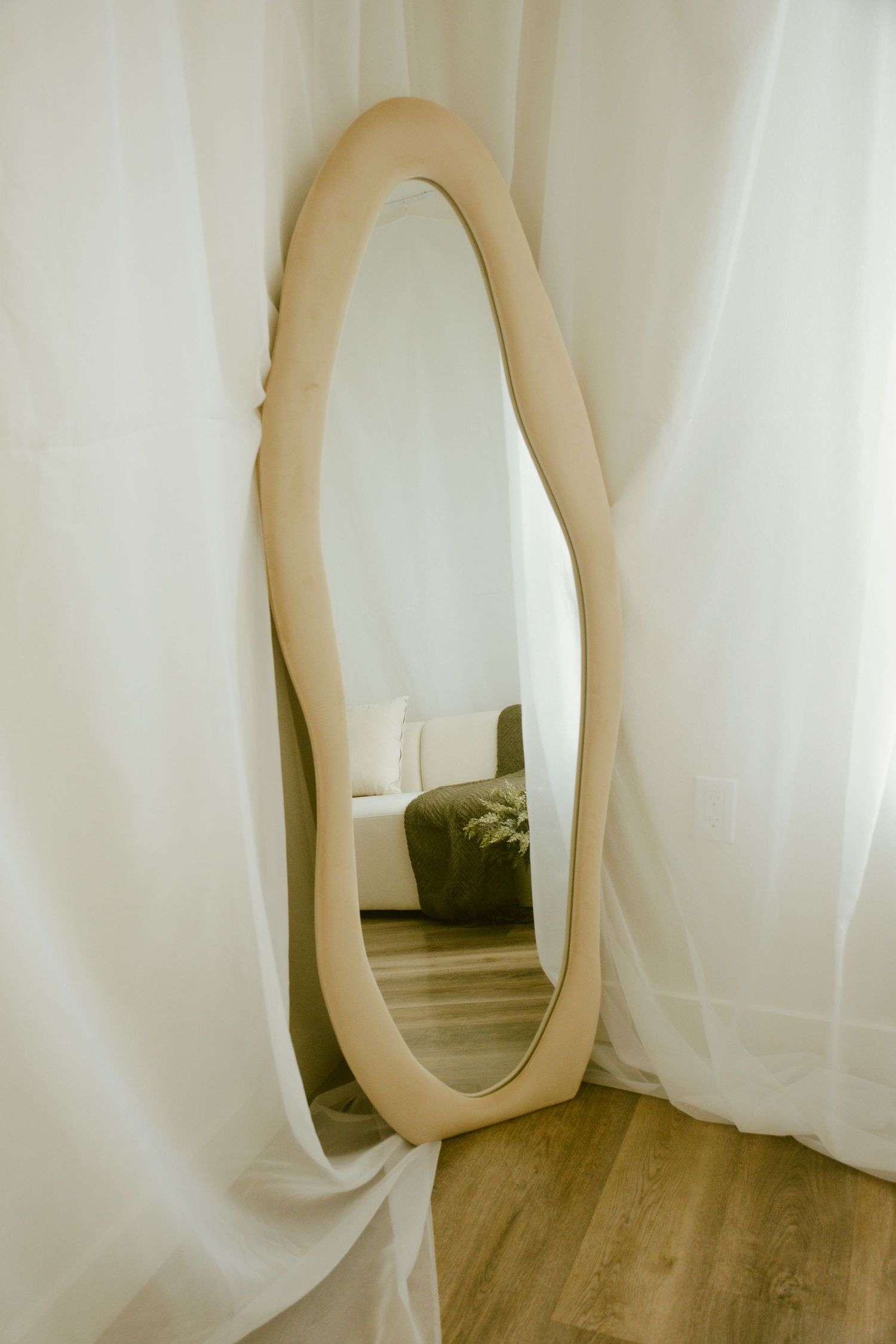 Curved beige mirror frame leaning against white curtained wall in minimal room setting.