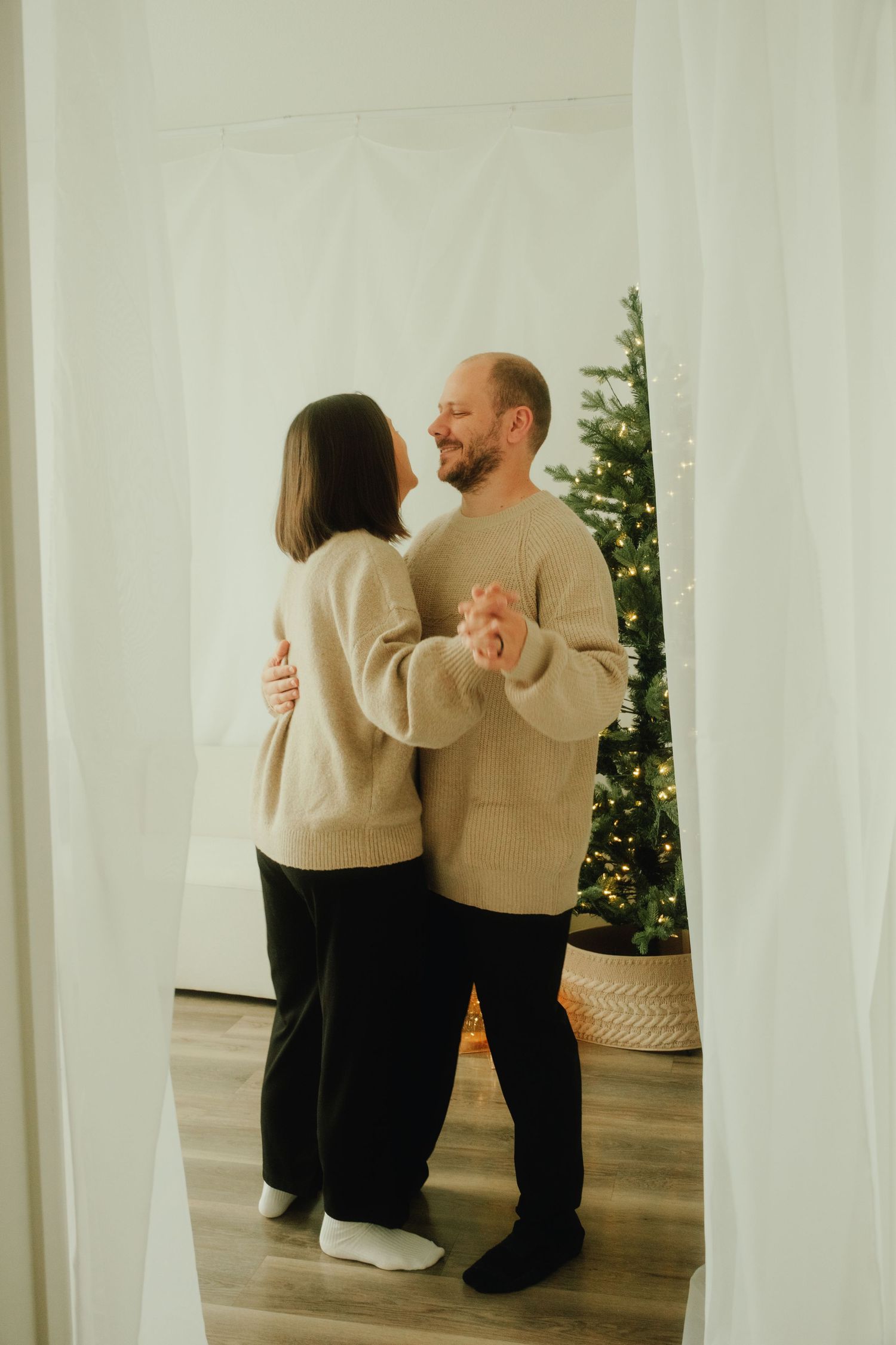 A couple in matching beige sweaters dancing together near a decorated Christmas tree.