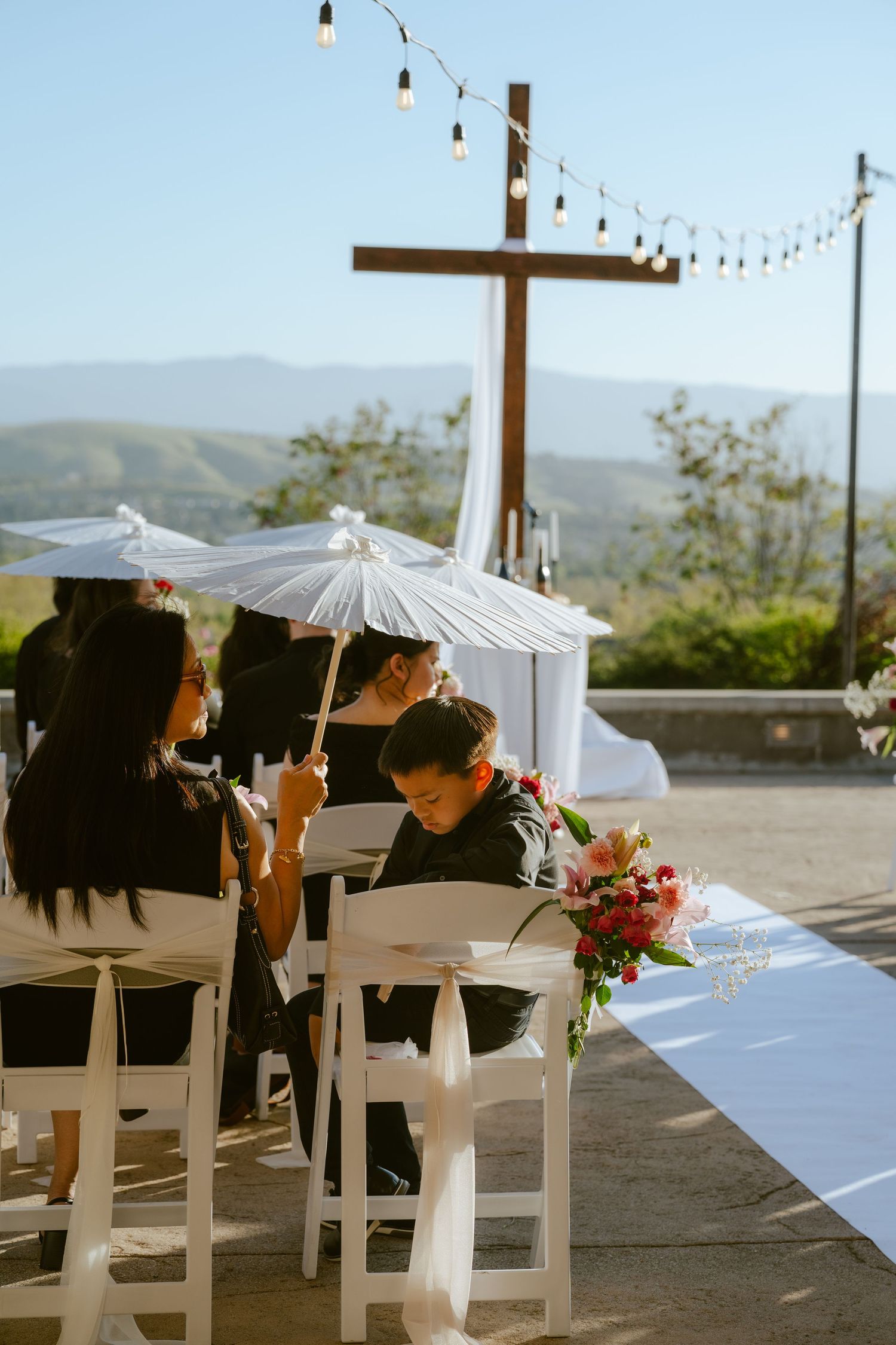 Outdoor wedding ceremony with guests holding white parasols, wooden cross altar, and mountain views.