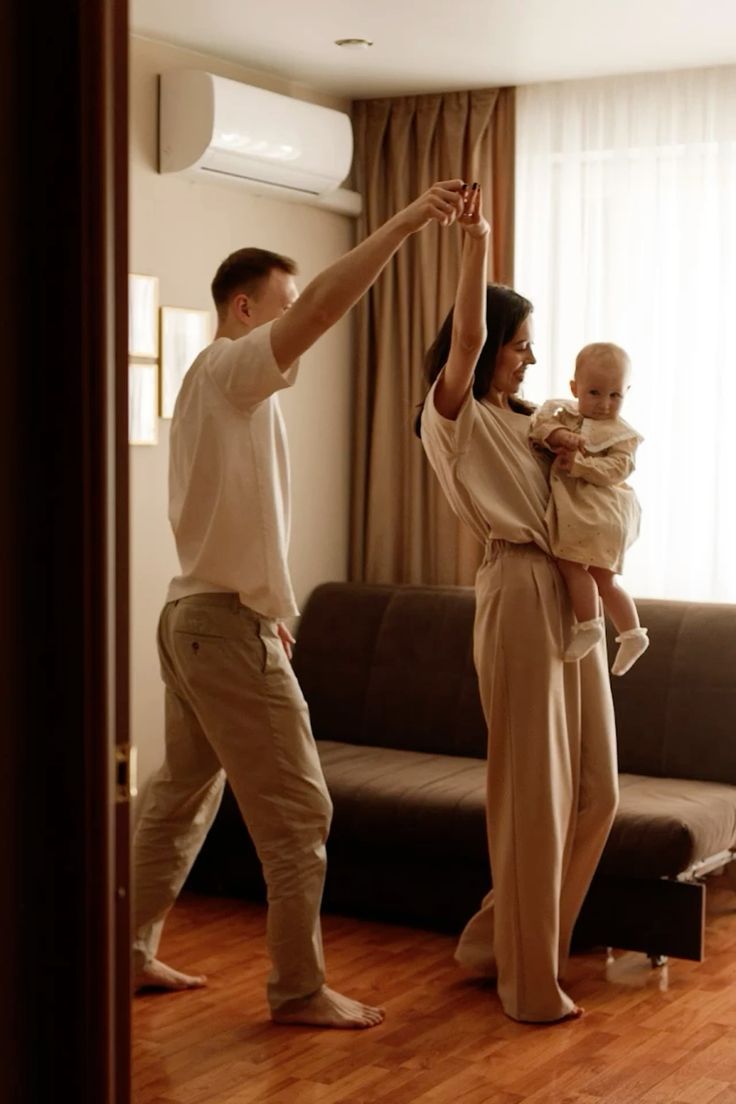 A family dances together in their living room with hardwood floors and natural lighting from the window.