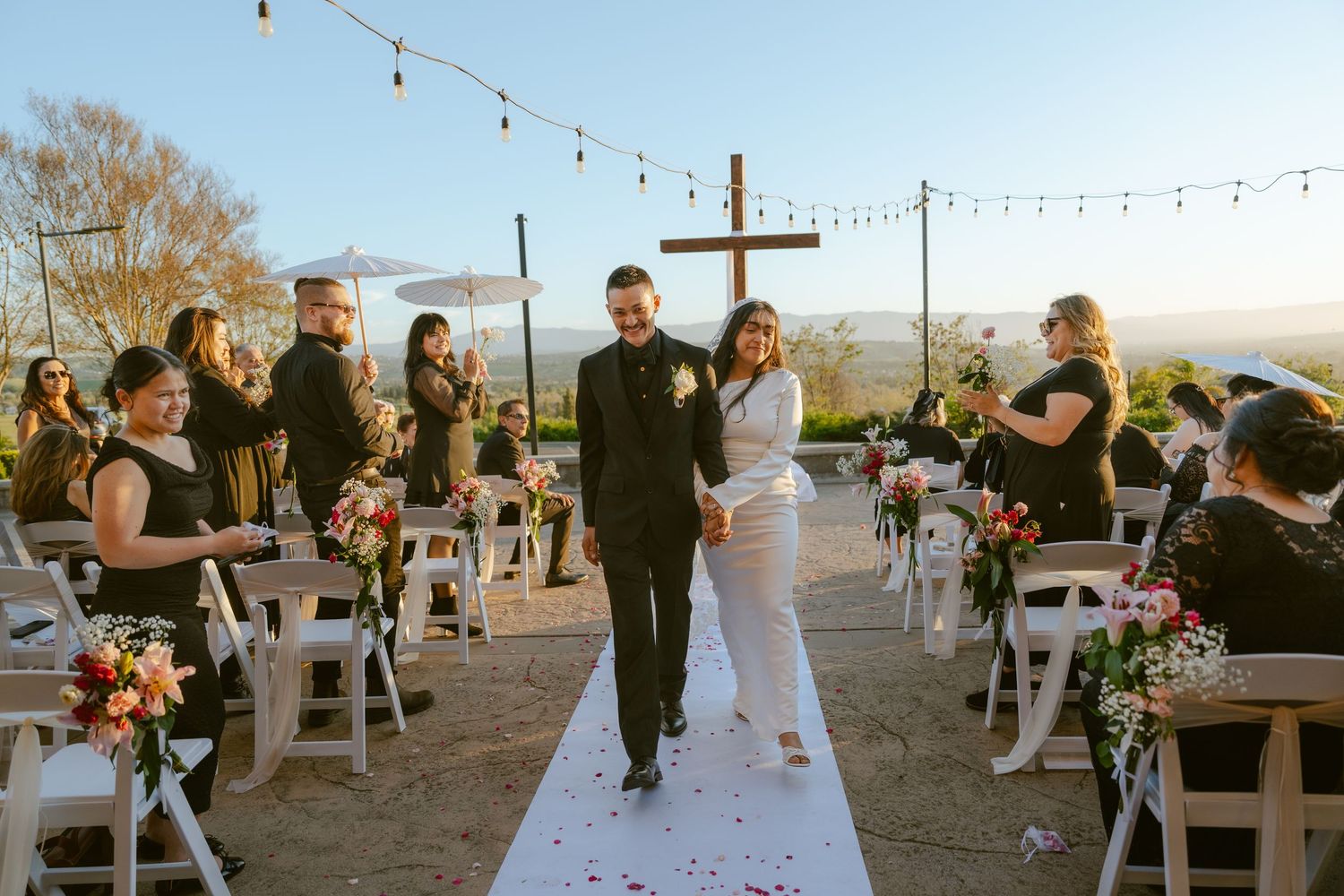 Newlywed couple walks down outdoor aisle at sunset wedding ceremony with cross backdrop and string lights.