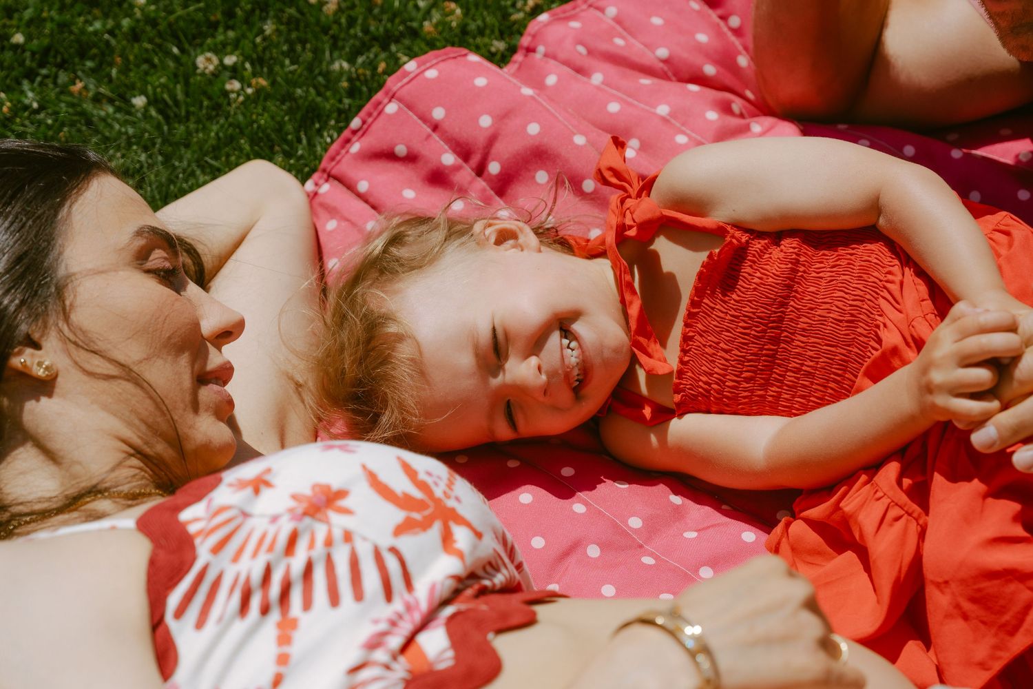 Mother and toddler girl in red dresses lying on a pink polka dot blanket in the grass, laughing together.
