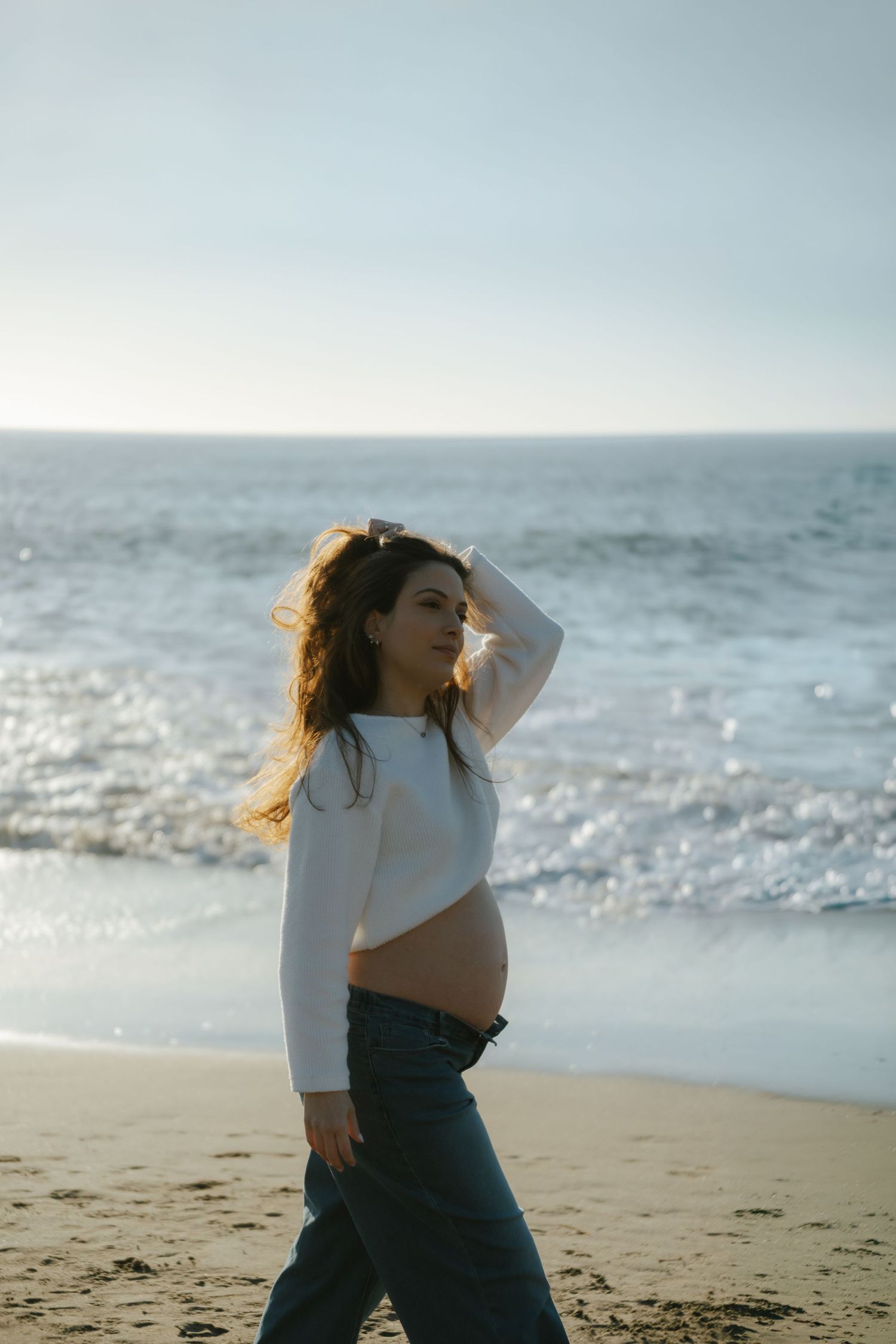 Pregnant woman in white crop top and jeans walking on a sandy beach with ocean waves in the background.