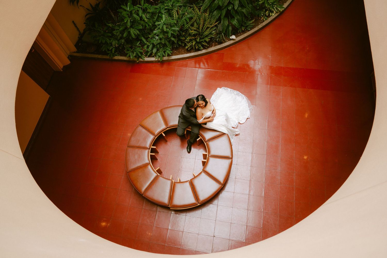 Aerial view of a wedding couple embracing on a circular staircase with red floors and lush greenery above.