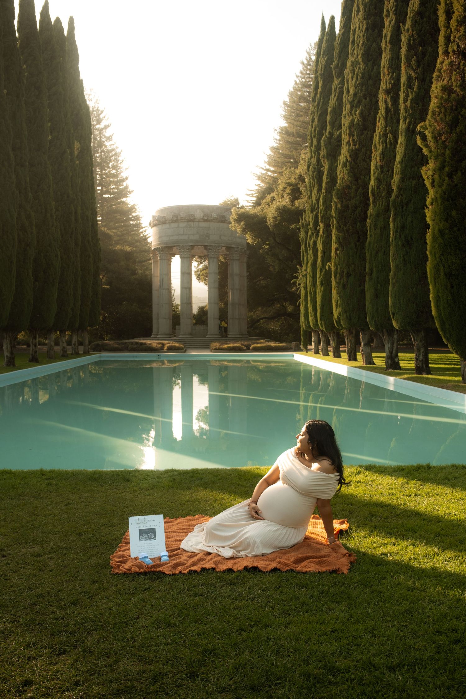 Pregnant woman in white dress sits by a serene reflecting pool surrounded by cypress trees and a classical stone pavilion.