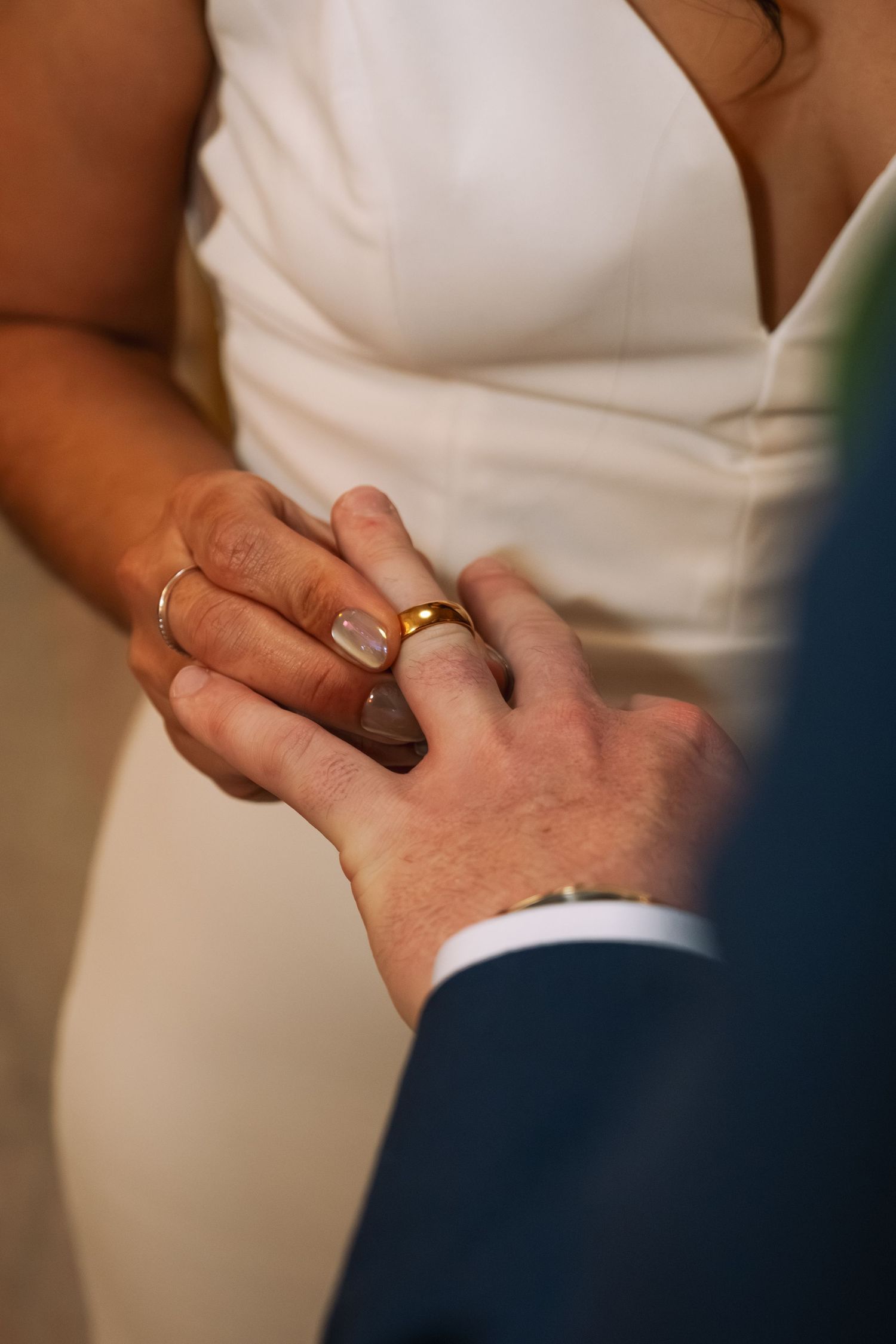 Close-up shot of hands exchanging wedding rings during a ceremony, with bride in white dress and groom in navy suit.