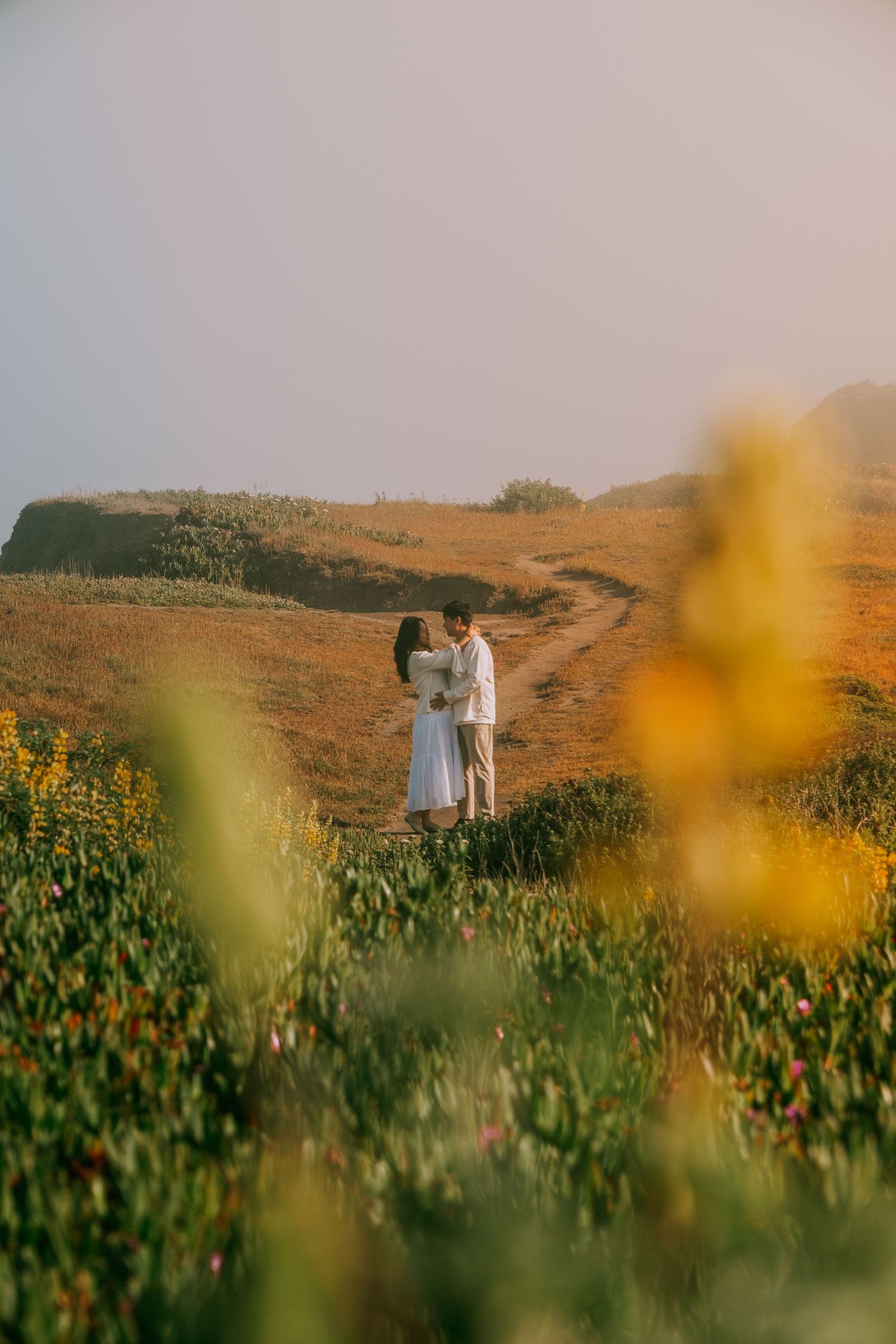 A couple walks through a golden meadow at sunset with wildflowers in the foreground.