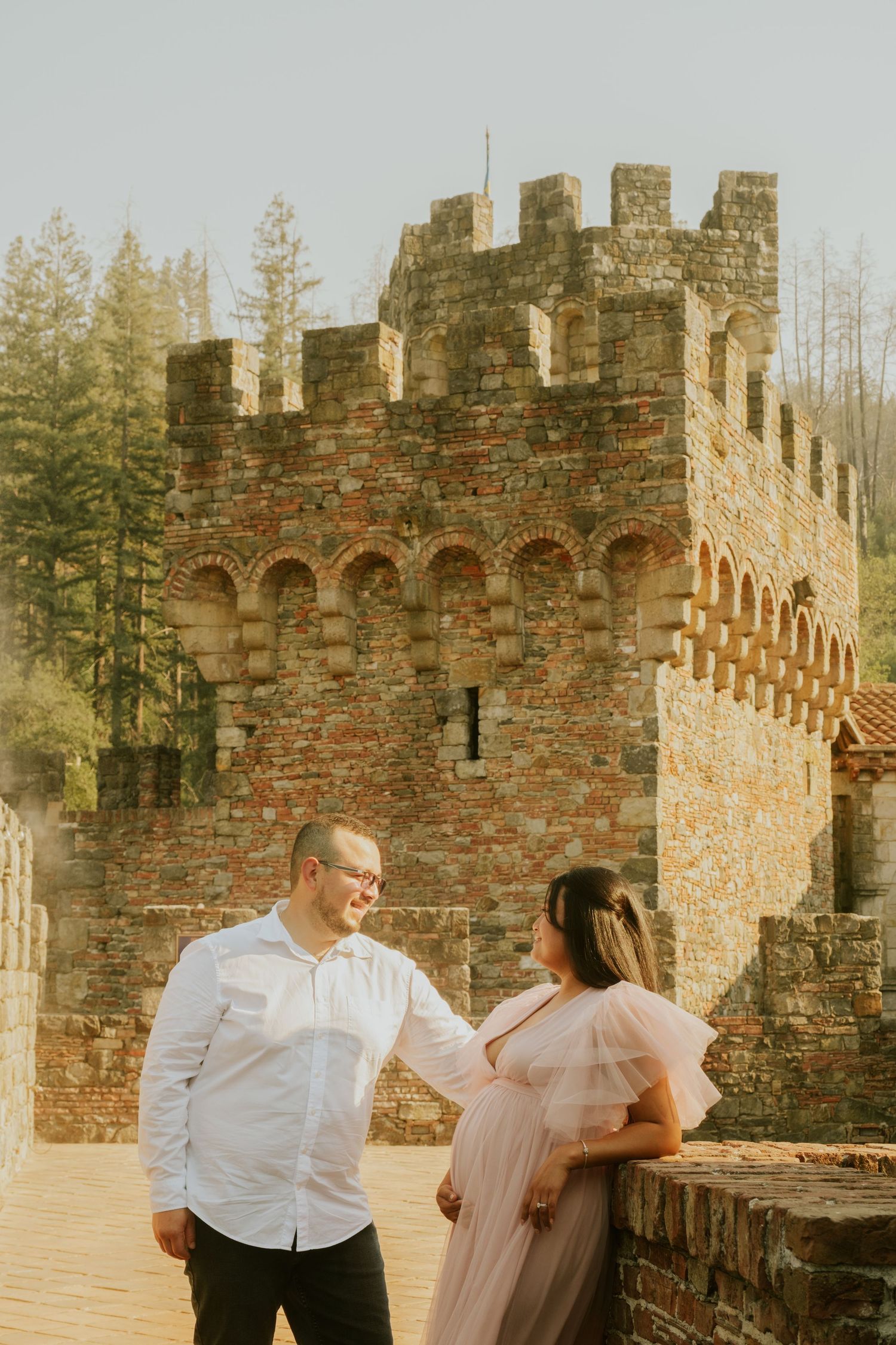 A couple poses playfully against ancient castle walls in warm sunlight.