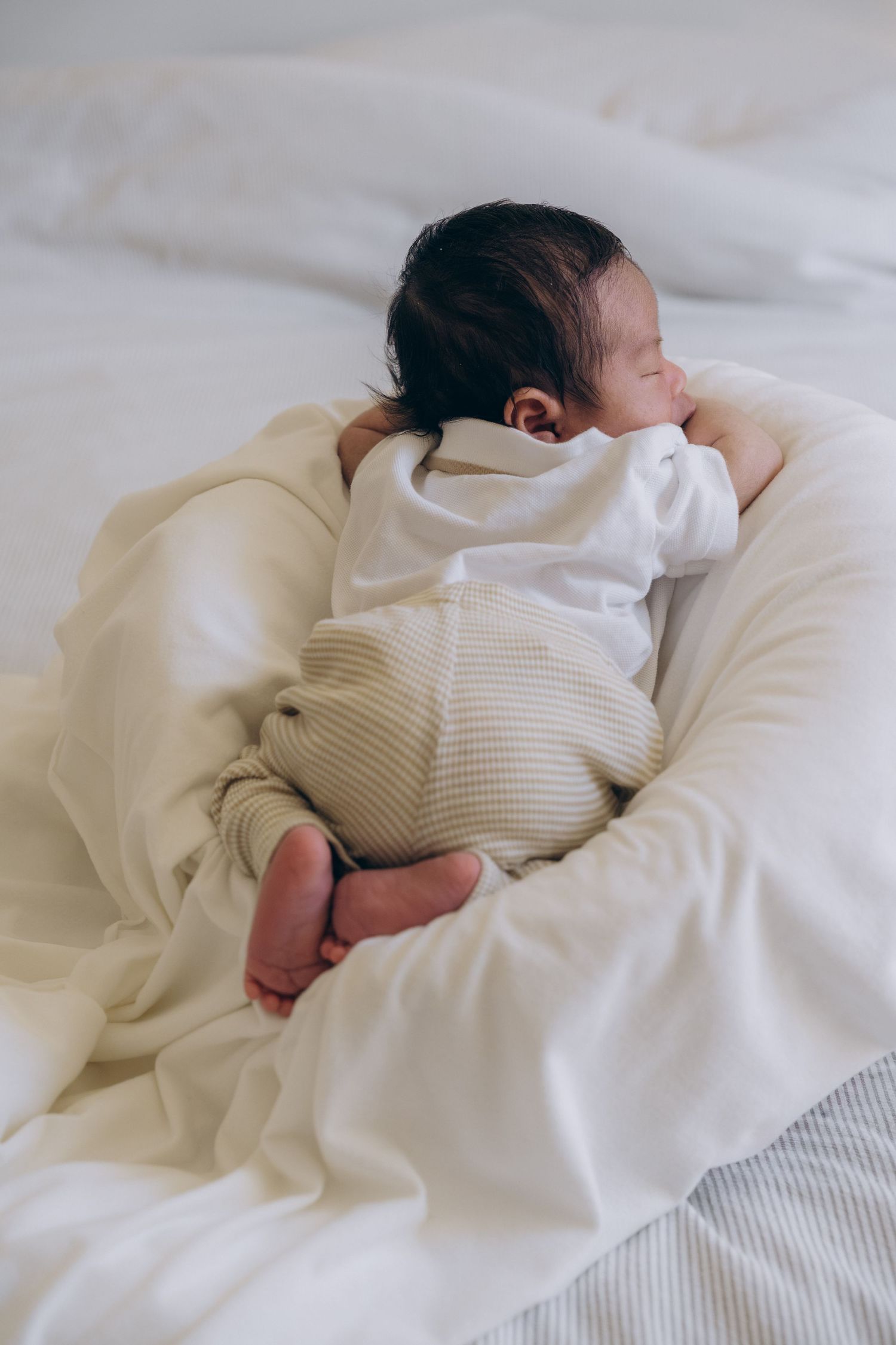 Sleeping newborn baby curled up on a white bed wrapped in cream and white soft blankets.