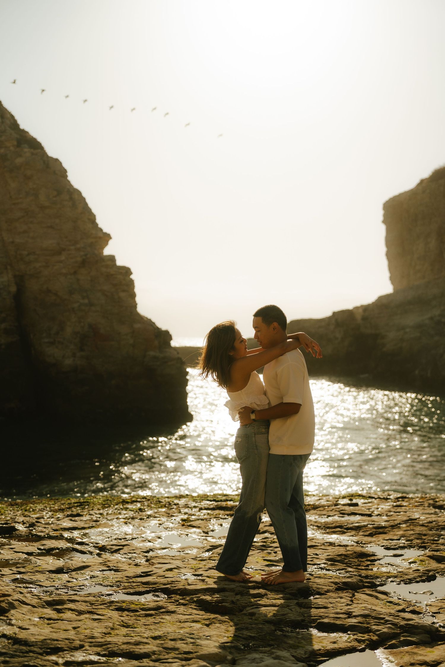 Couple embracing romantically on rocky coastal shore at golden hour, framed by dramatic cliffs with sunlit water behind them.