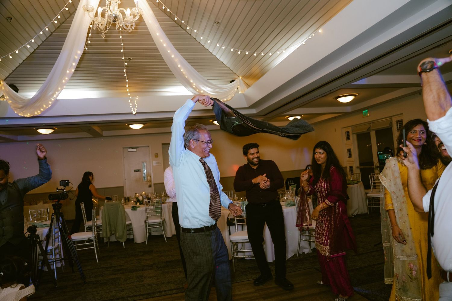 Guests dancing joyfully at an elegant indoor wedding reception with fairy lights and draped ceiling decor.