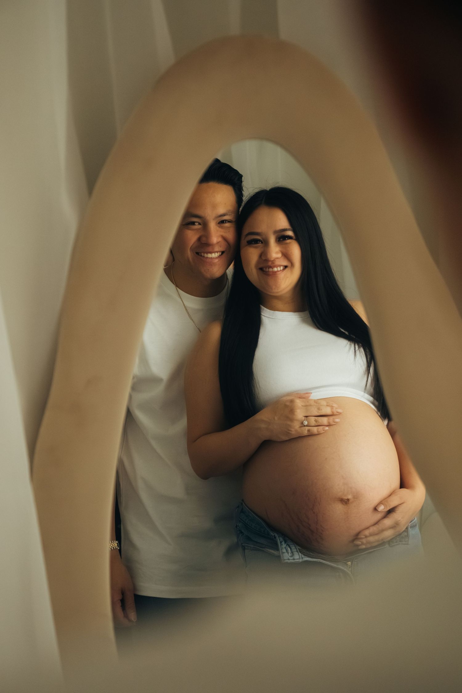 Artistic maternity photo through curved frame showing expecting couple in white against neutral background.