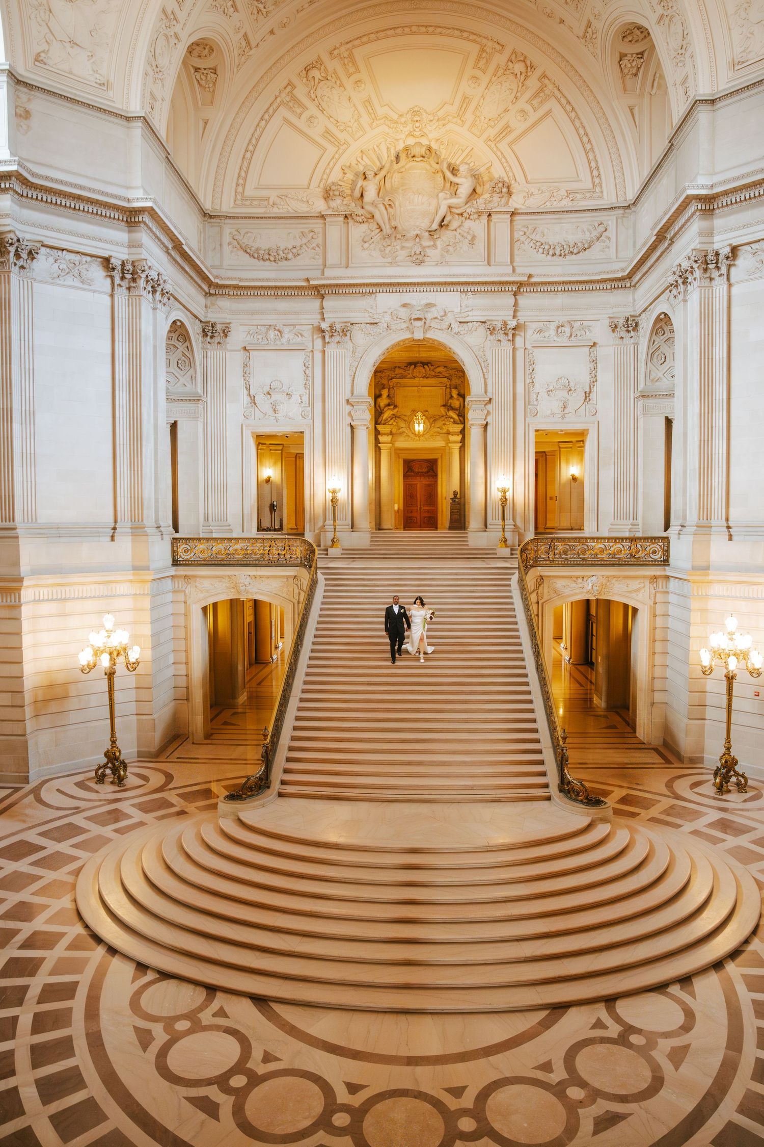 Bride and groom stand on grand marble staircase inside San Francisco City Hall's ornate Beaux-Arts rotunda.