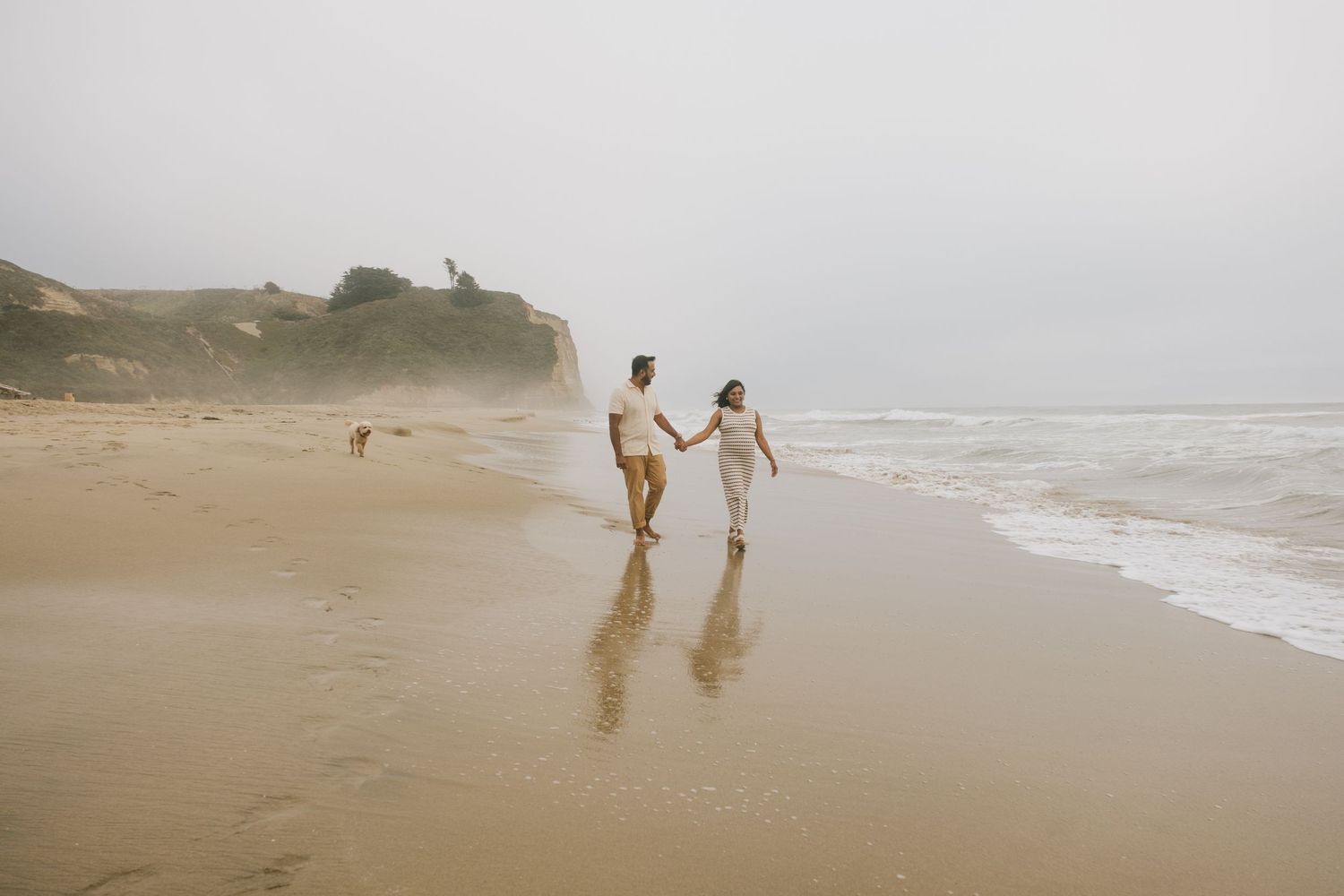 Couple walks hand in hand along misty beach shoreline.