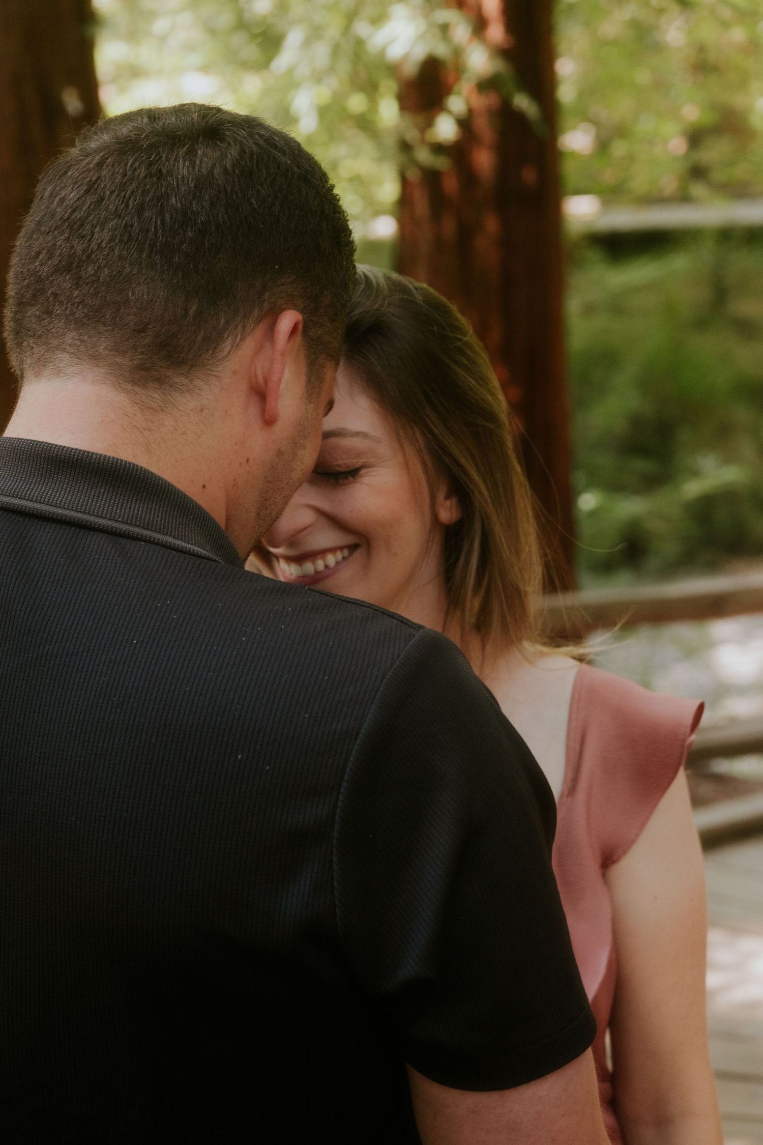 Romantic couple sharing intimate moments on a wooden boardwalk surrounded by towering redwood trees.