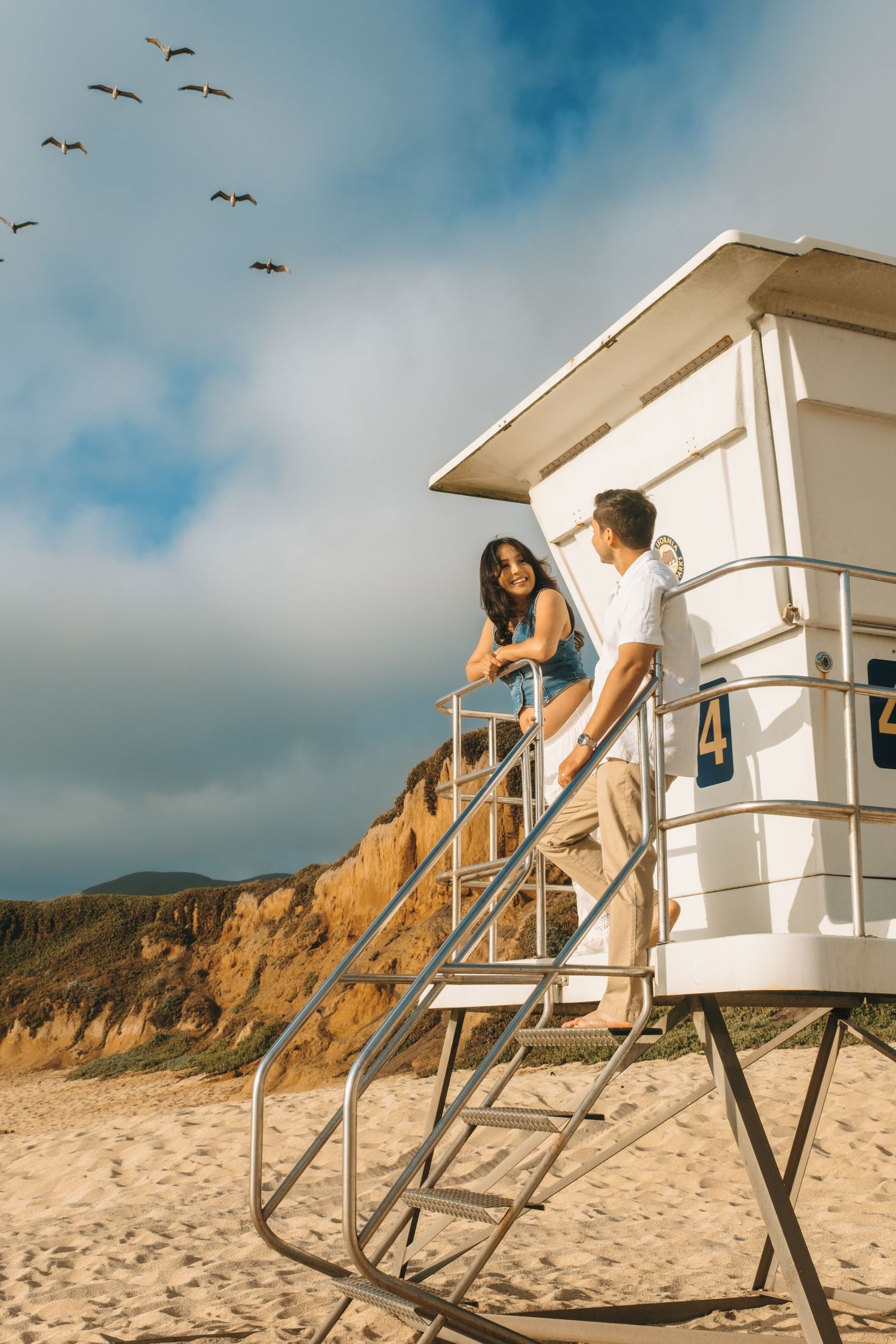A couple poses playfully on a lifeguard tower at the beach with seagulls flying overhead.