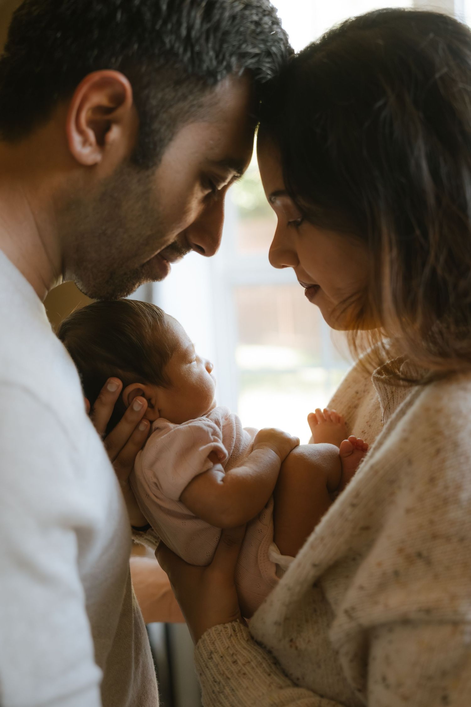 New parents tenderly cradle their sleeping newborn baby, foreheads touching in an intimate family portrait.