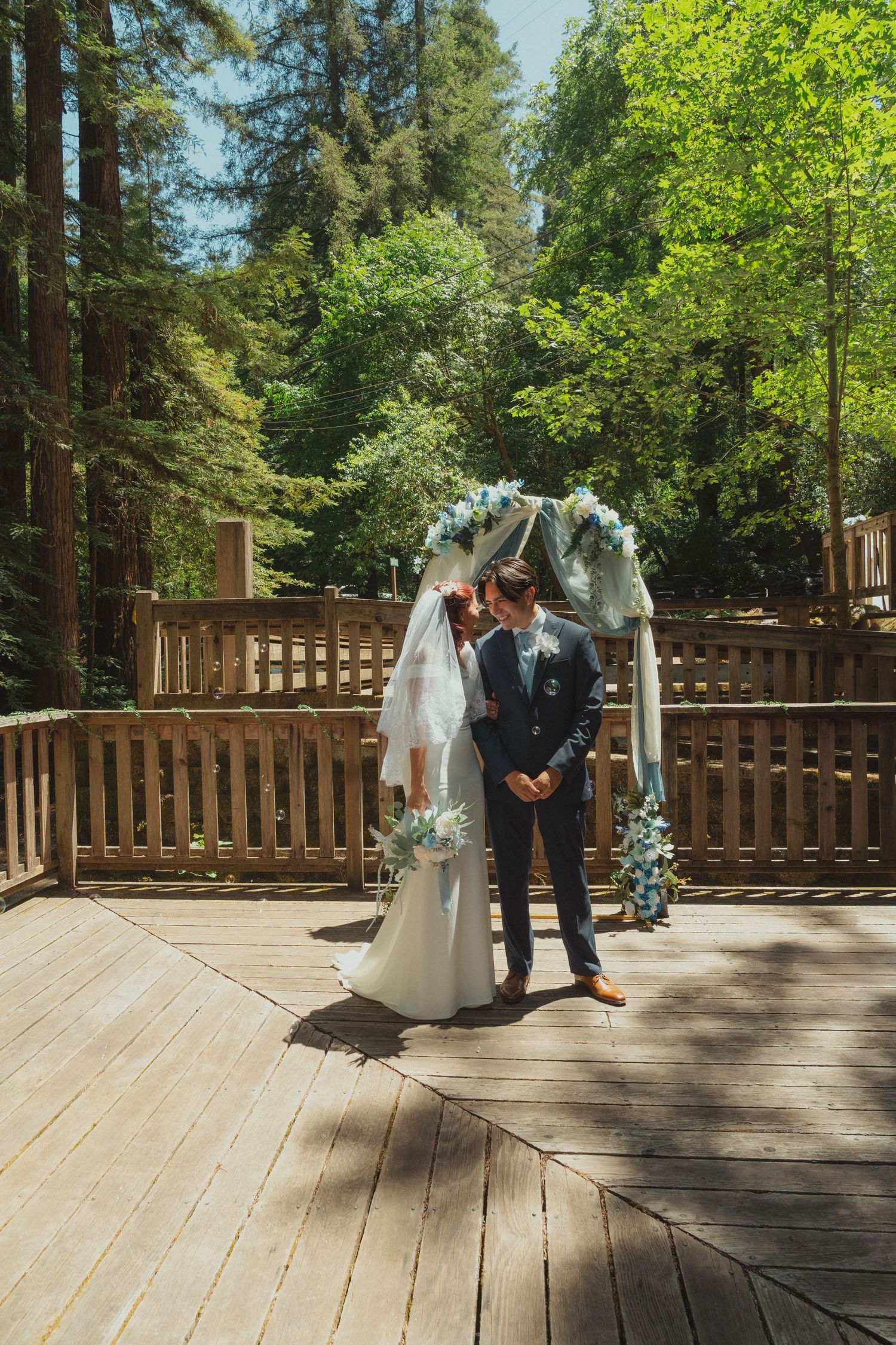Bride and groom kissing under a floral arch on a wooden deck surrounded by lush green trees at an outdoor wedding.