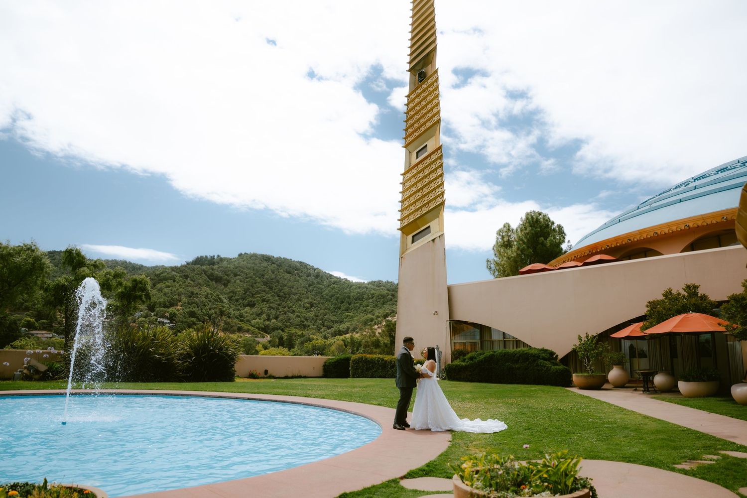 Bride and groom stand by a fountain with mountain views at an elegant outdoor wedding venue.