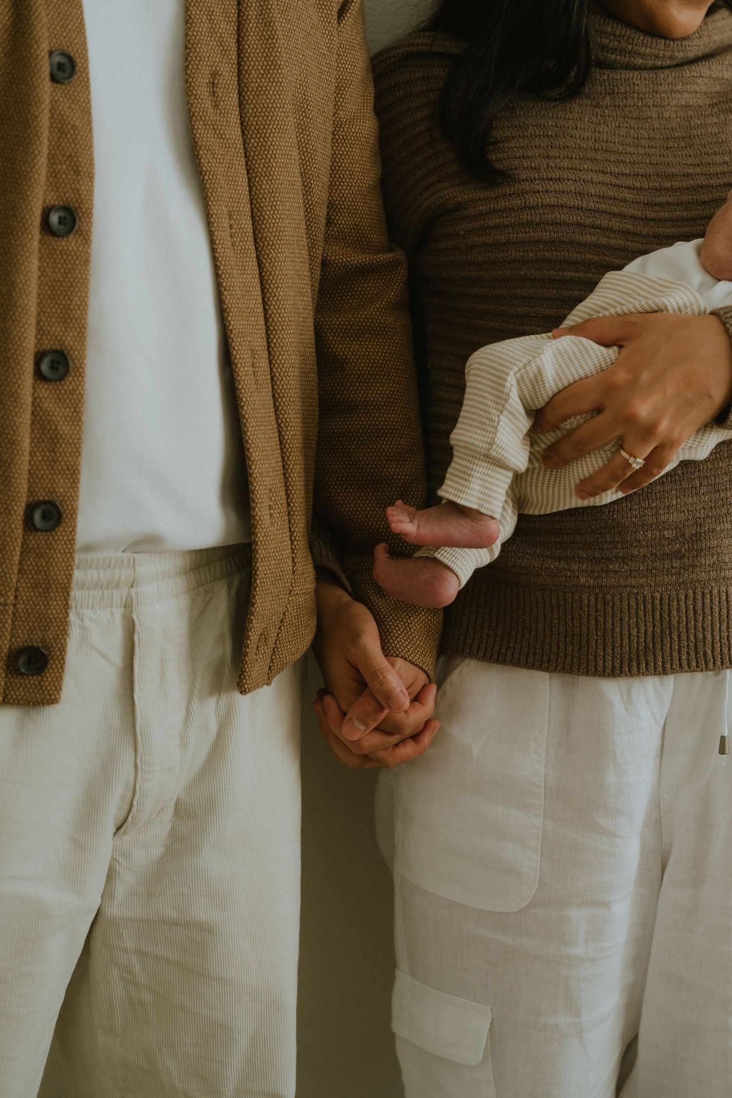 A couple in neutral tones holds hands while cradling a newborn baby, capturing a tender family moment.