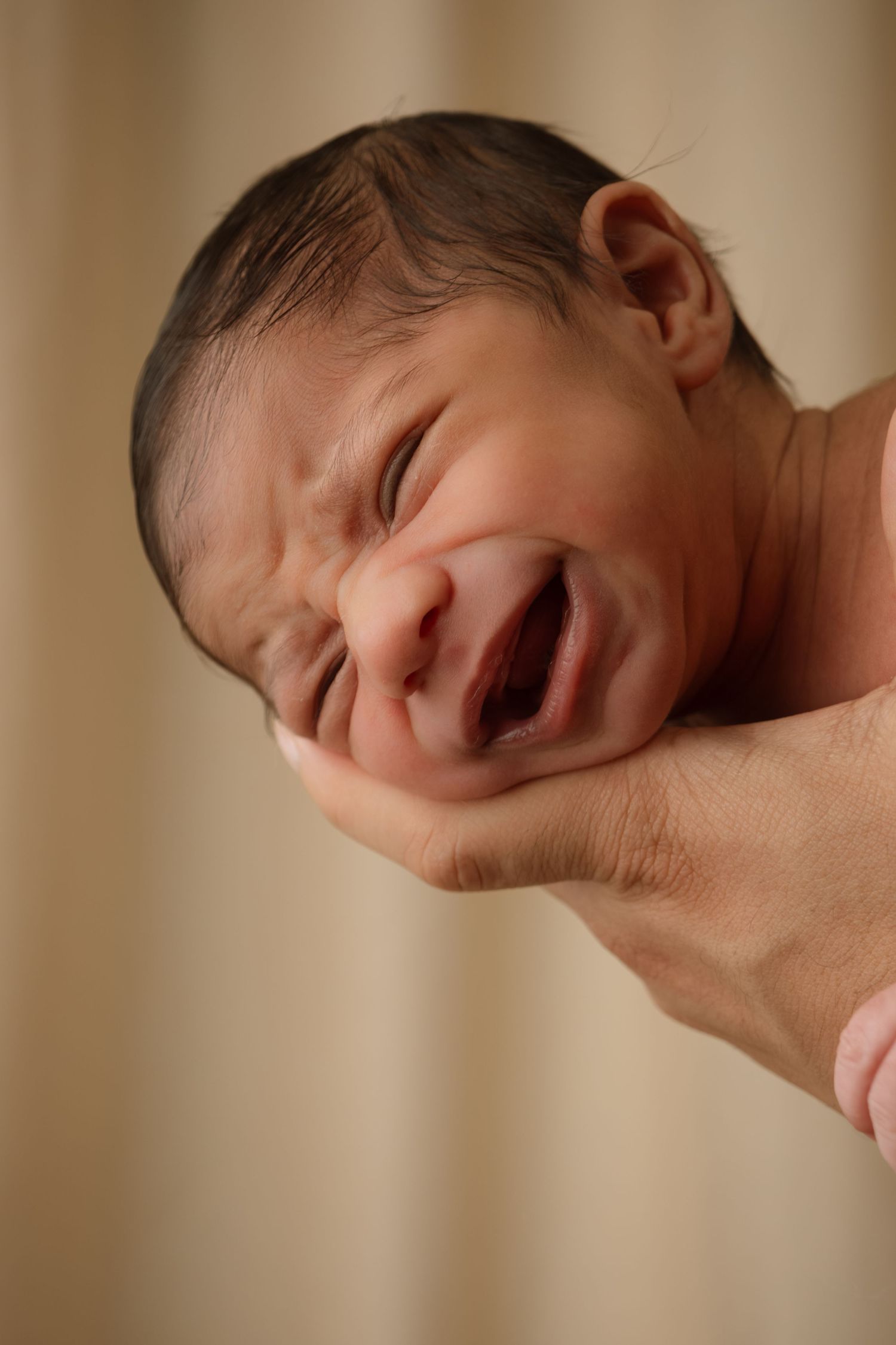 Side profile of newborn baby with expressive face against warm brown background.