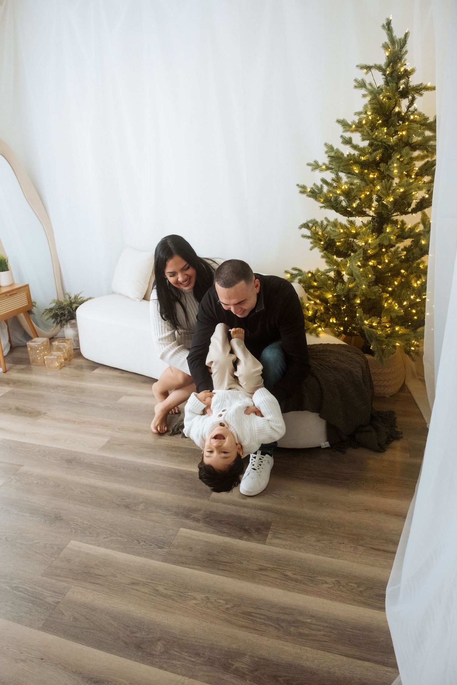Family sharing playful moment on hardwood floor by illuminated Christmas tree in bright white room.