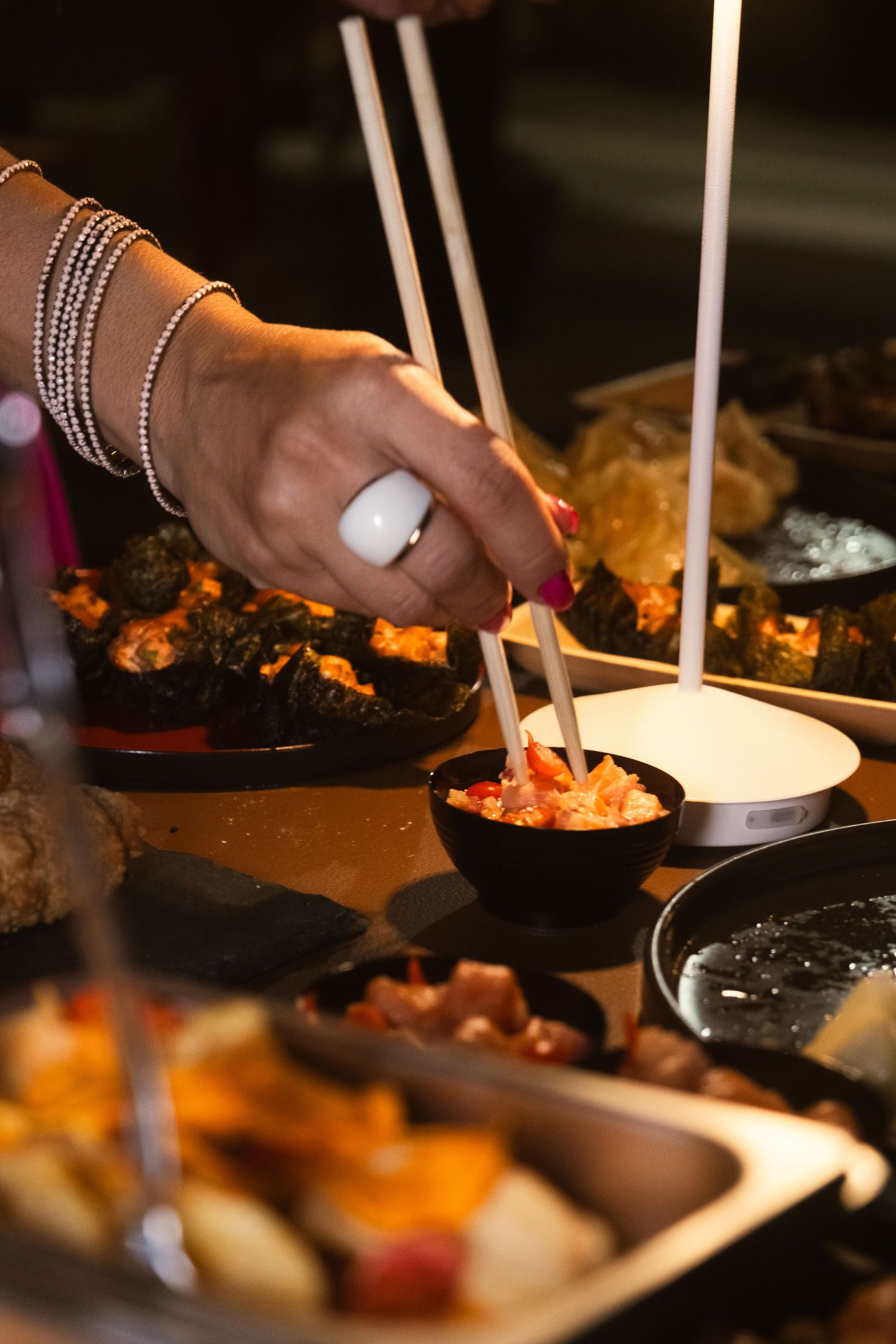 Hand with white nail polish and bracelets using chopsticks to serve Asian cuisine.