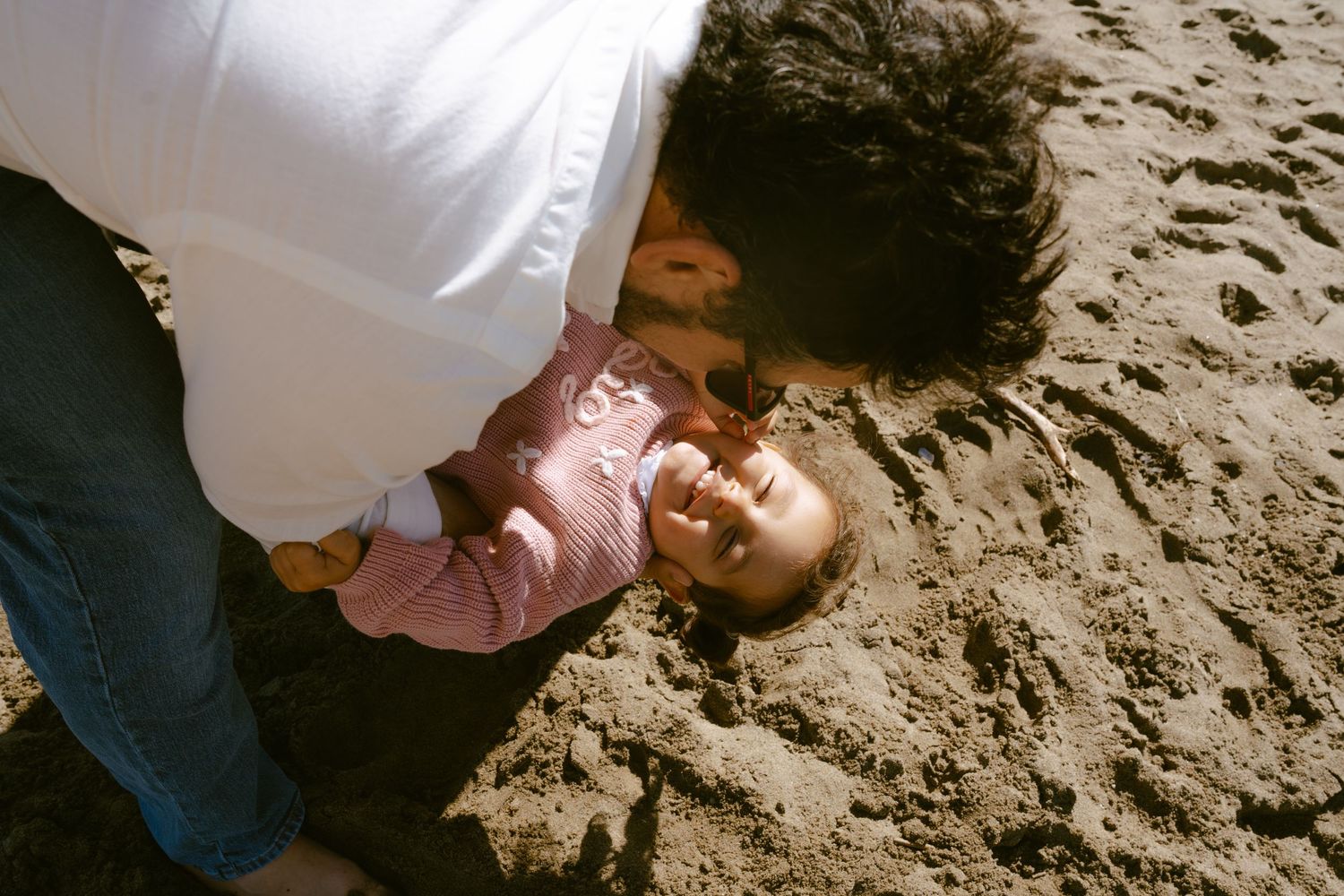 Parent lovingly leans over smiling baby lying on sandy beach, sharing a joyful moment outdoors.