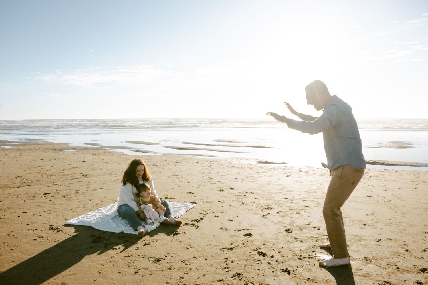Family enjoying a sunny beach day, with a woman sitting on a blanket and a man gesturing playfully near the ocean.