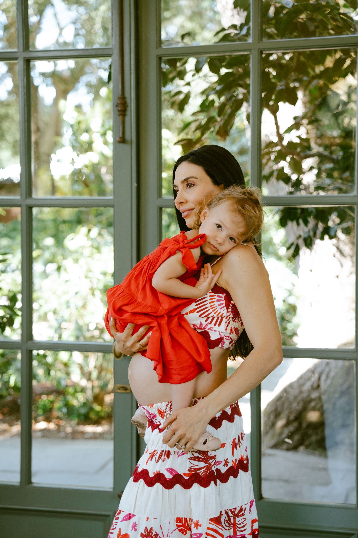 Pregnant woman in floral dress holds toddler in red outfit near glass doors with green foliage backdrop.