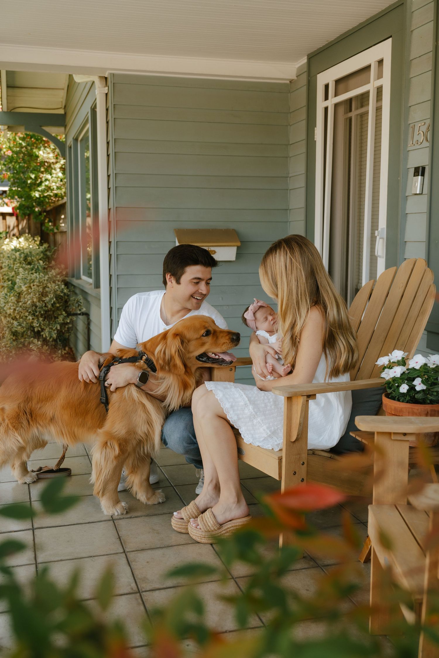 Young couple with golden retriever dog and newborn baby relaxing on home front porch in wooden chairs.