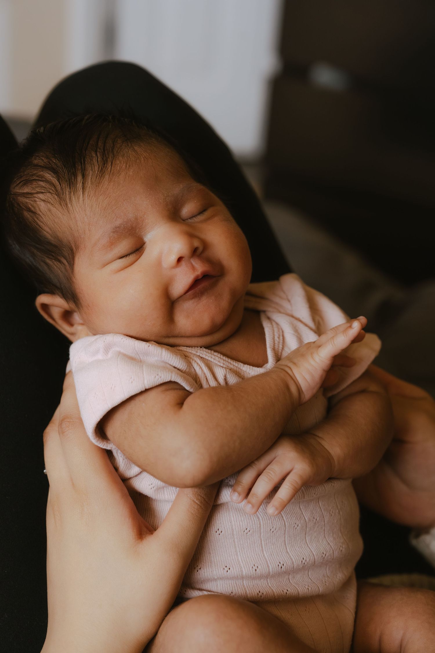 Newborn baby in pink onesie smiling with eyes closed while being cradled in an adult's hands.