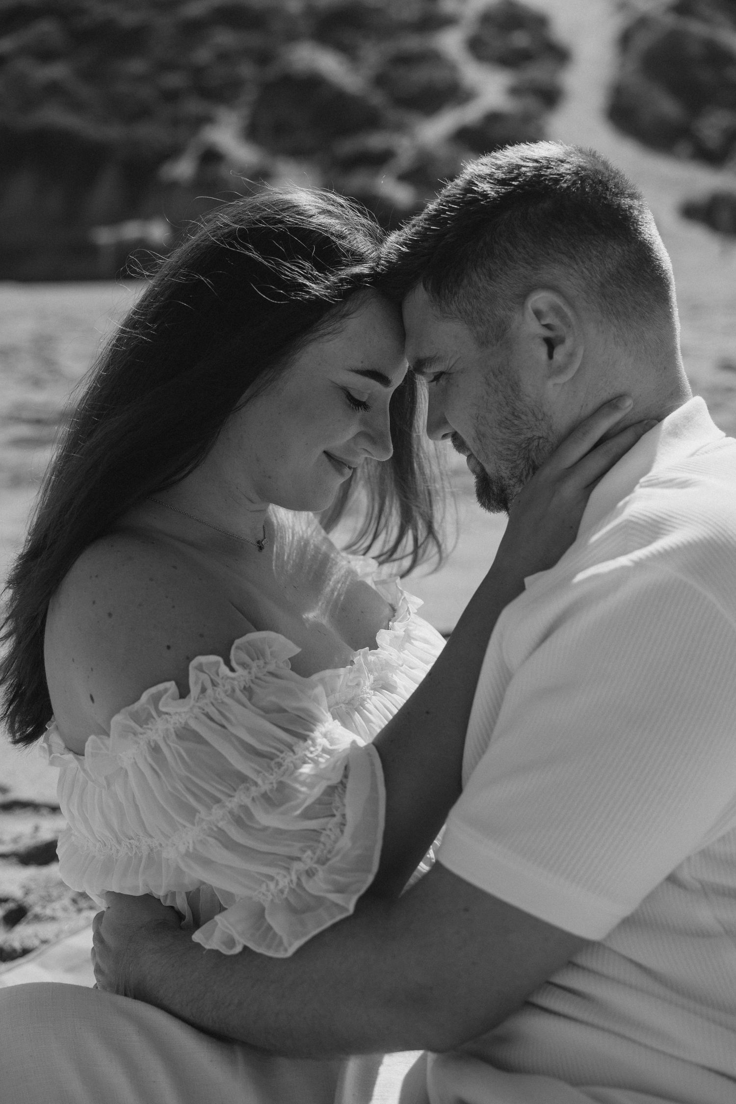 Black and white photo of romantic couple embracing forehead to forehead, woman in ruffled white dress outdoors.