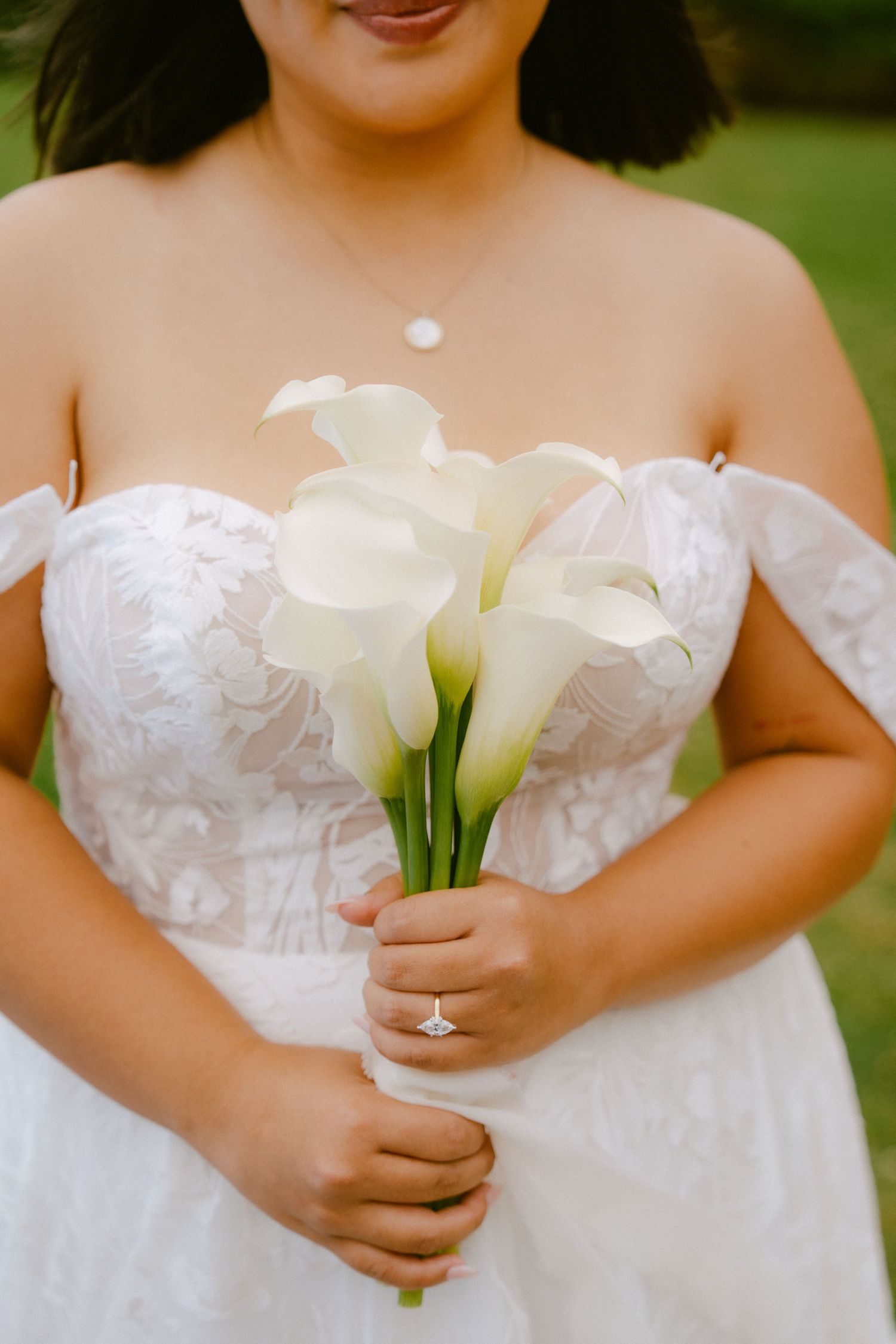 Bride in off-shoulder lace gown holds white calla lily bouquet, diamond ring visible, green background.