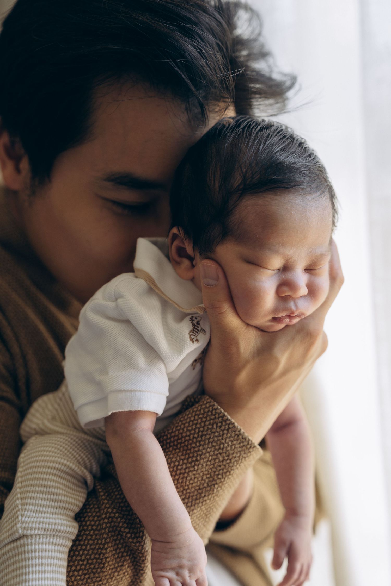 A young child tenderly kisses a sleeping newborn baby held gently in their arms near a window.
