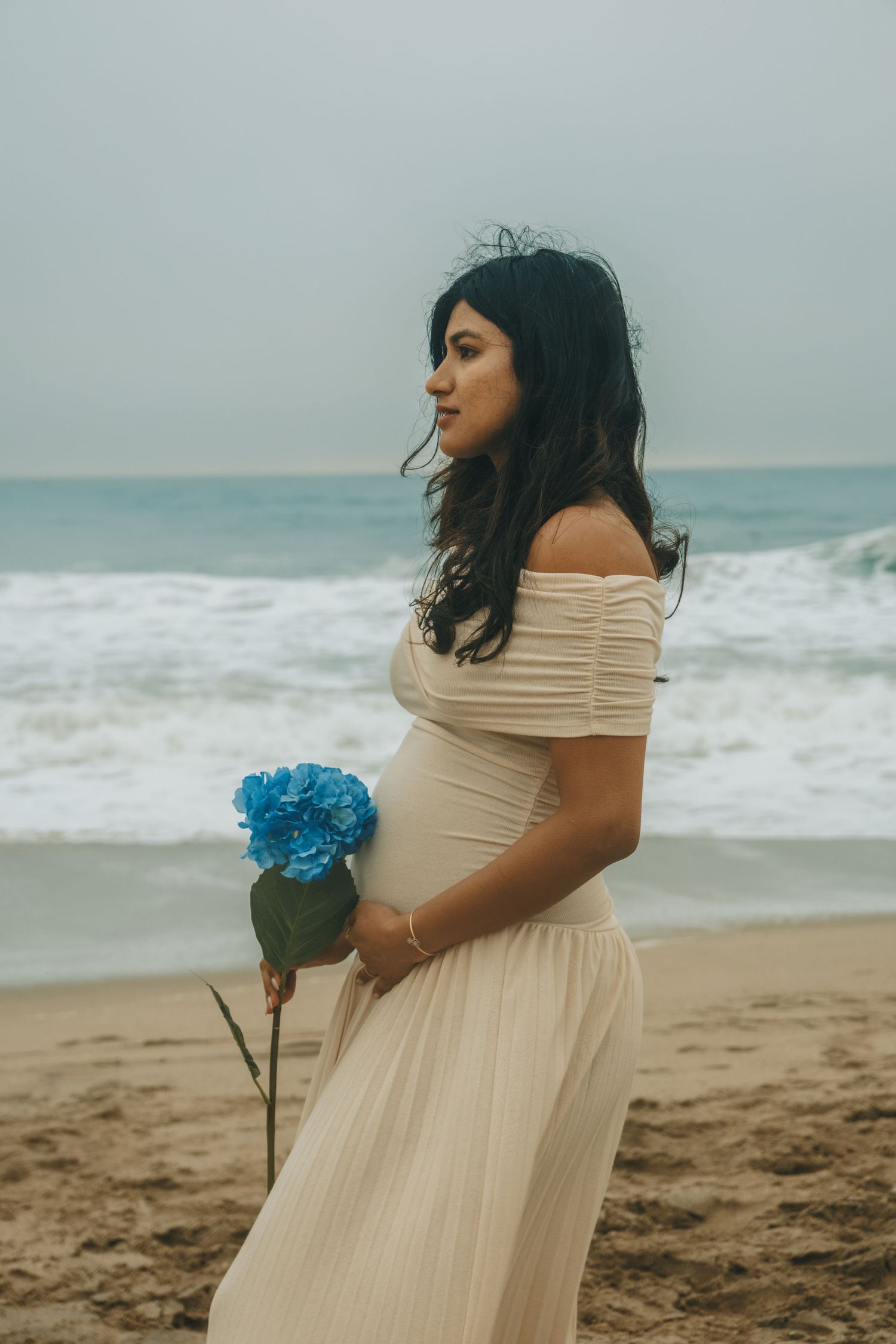 A person in an off-shoulder cream dress holds blue flowers on a beach.