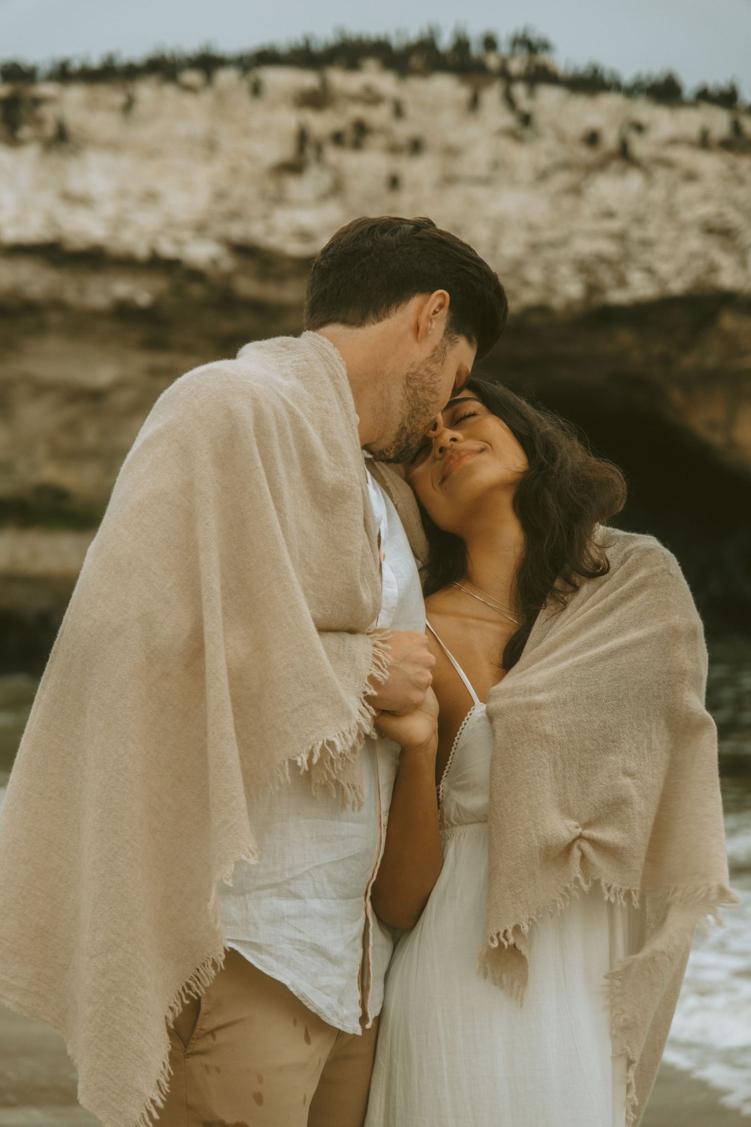 Couple wrapped in blankets share intimate moment on beach at sunset.