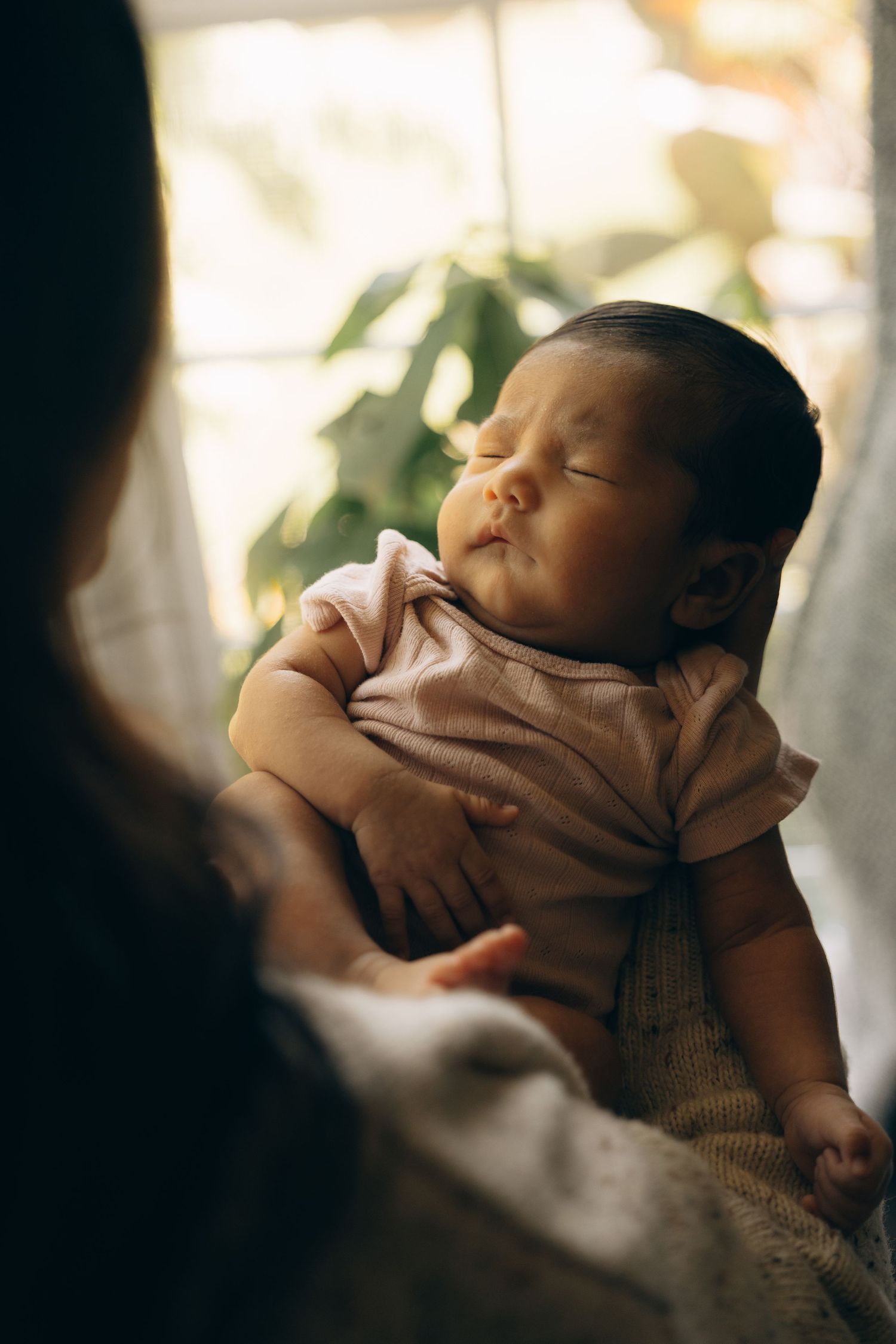 Peaceful newborn baby sleeping in mother's arms wearing a pink onesie in soft natural light.