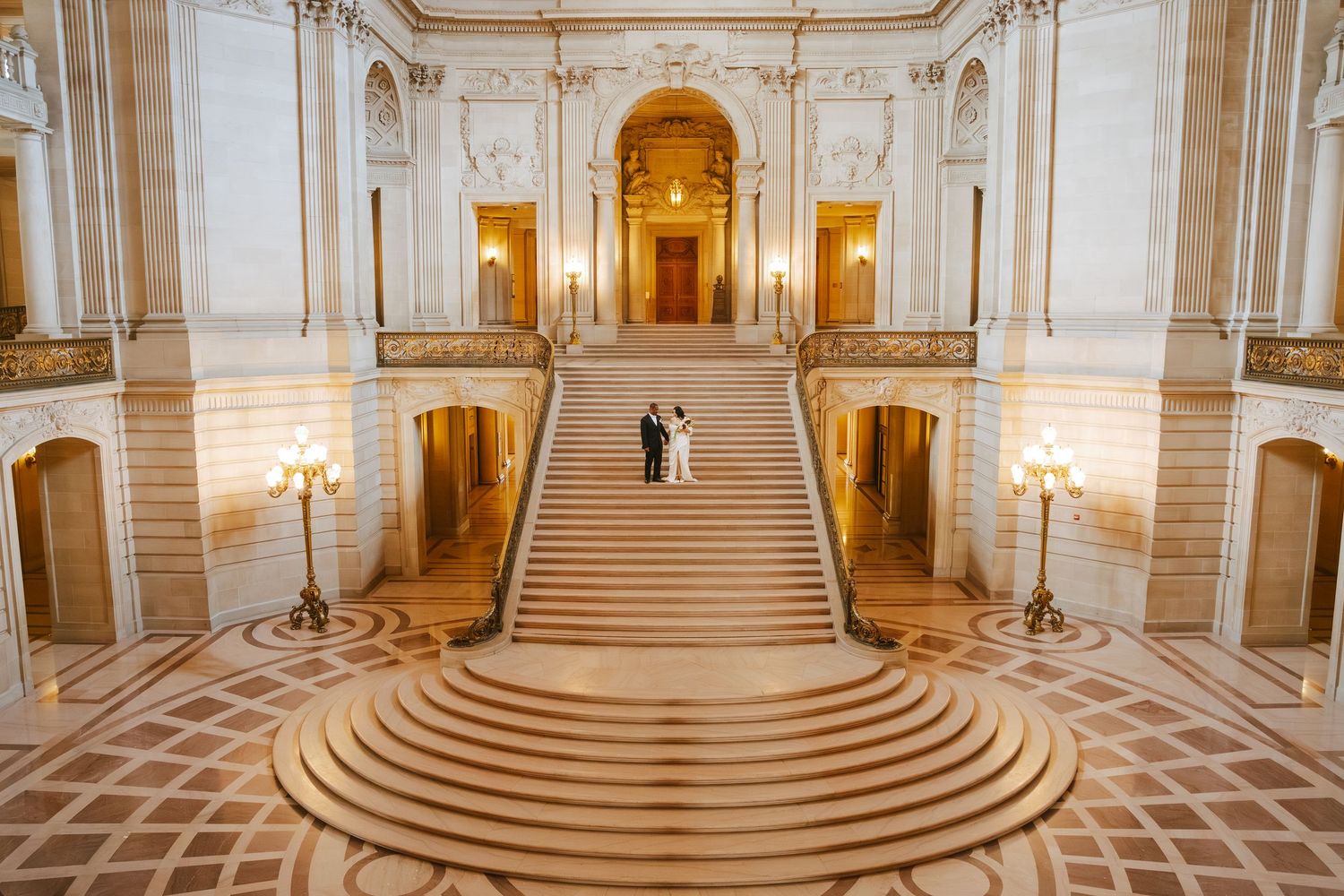 Couple poses on grand staircase of San Francisco City Hall, surrounded by ornate gold architecture and elegant marble floors.