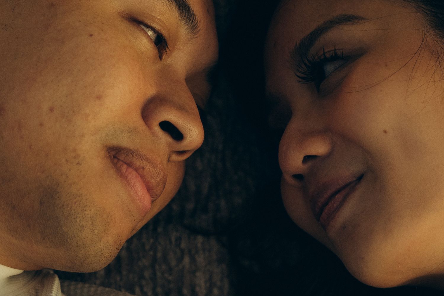 Close-up of a young couple lying face to face, smiling intimately in warm, soft lighting.