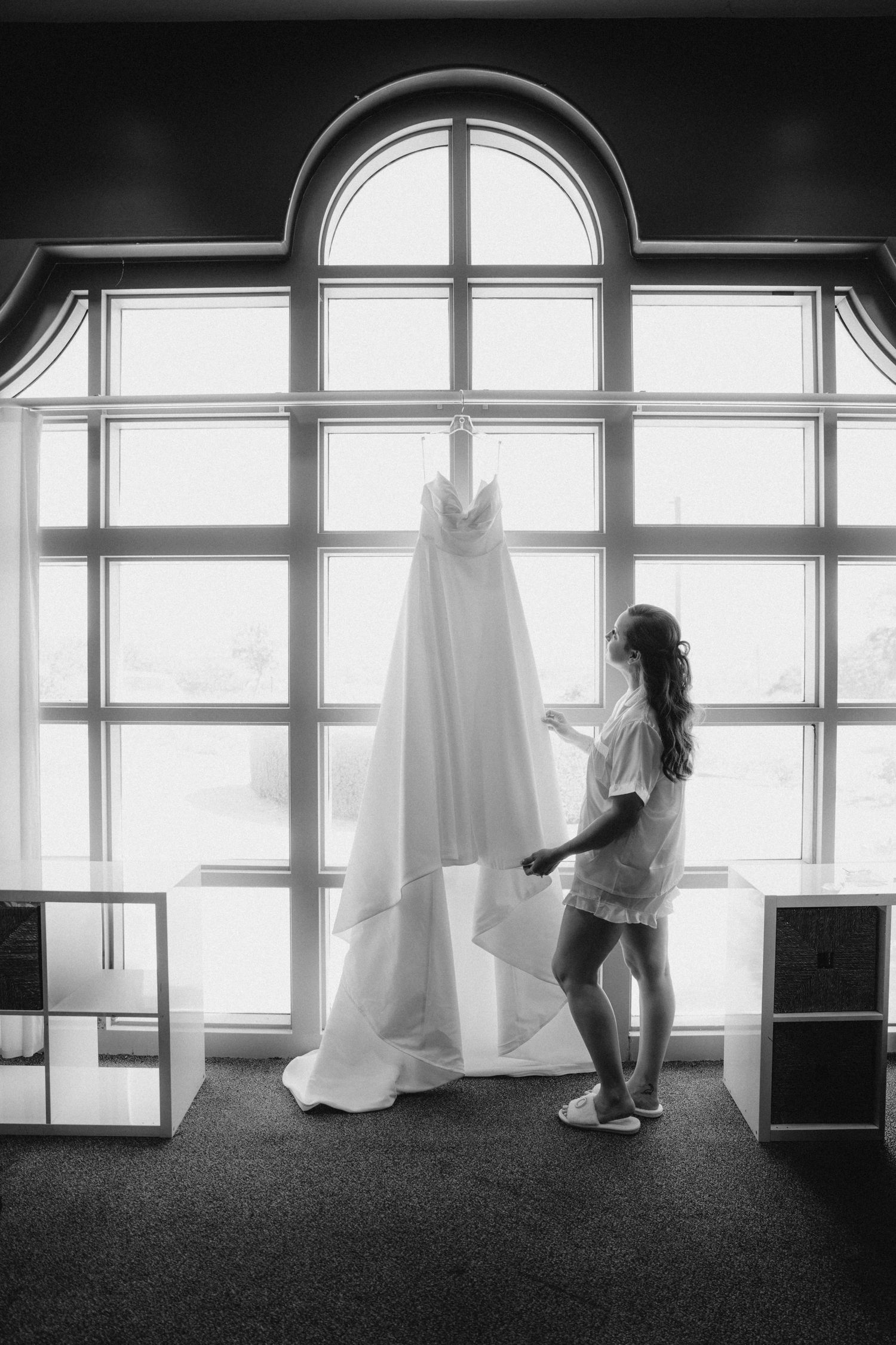 Black and white photo of a bride admiring her wedding gown hanging by a large arched window before the ceremony.