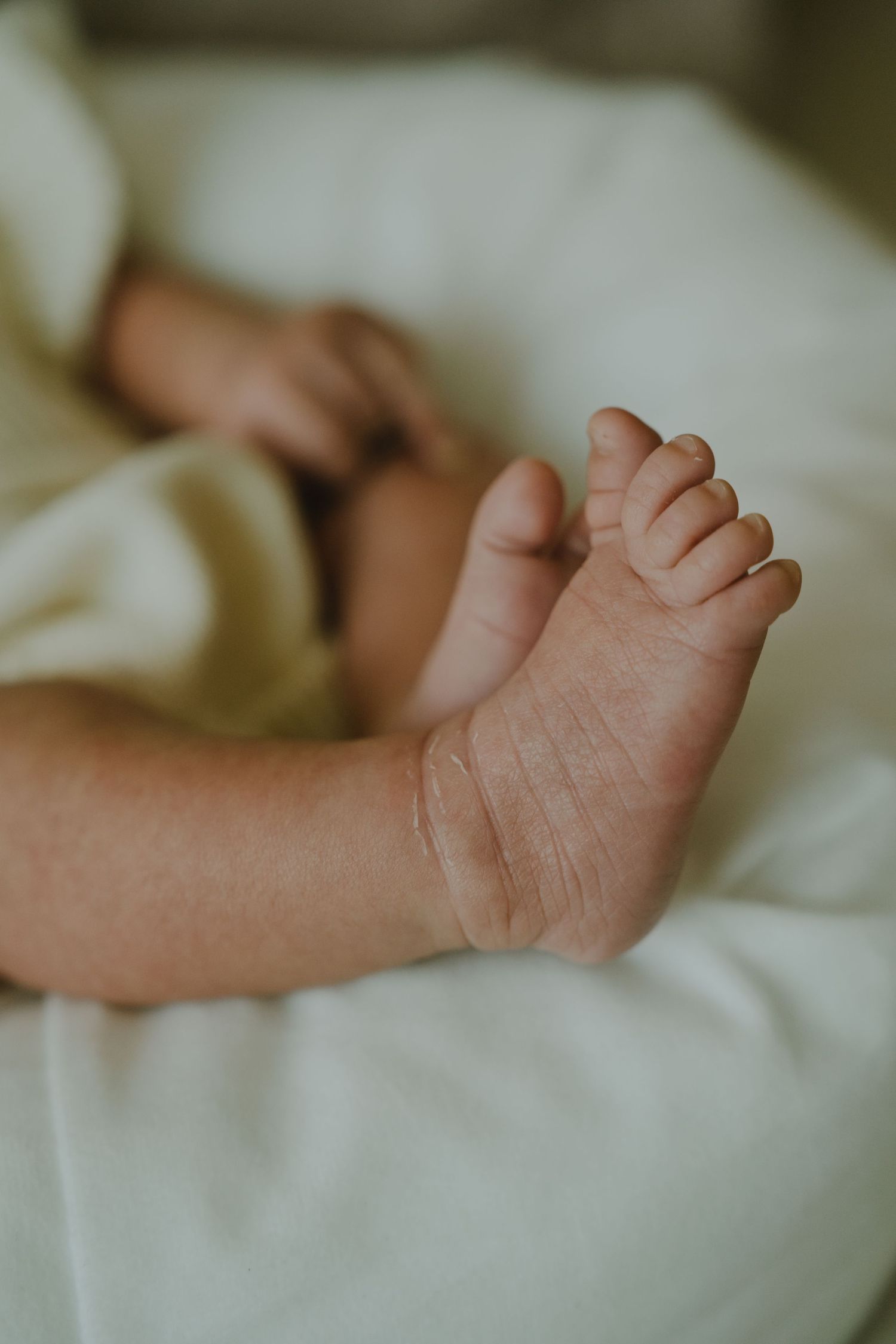 Close-up of tiny newborn baby feet resting on a soft white blanket, with delicate toes and chubby ankles visible.