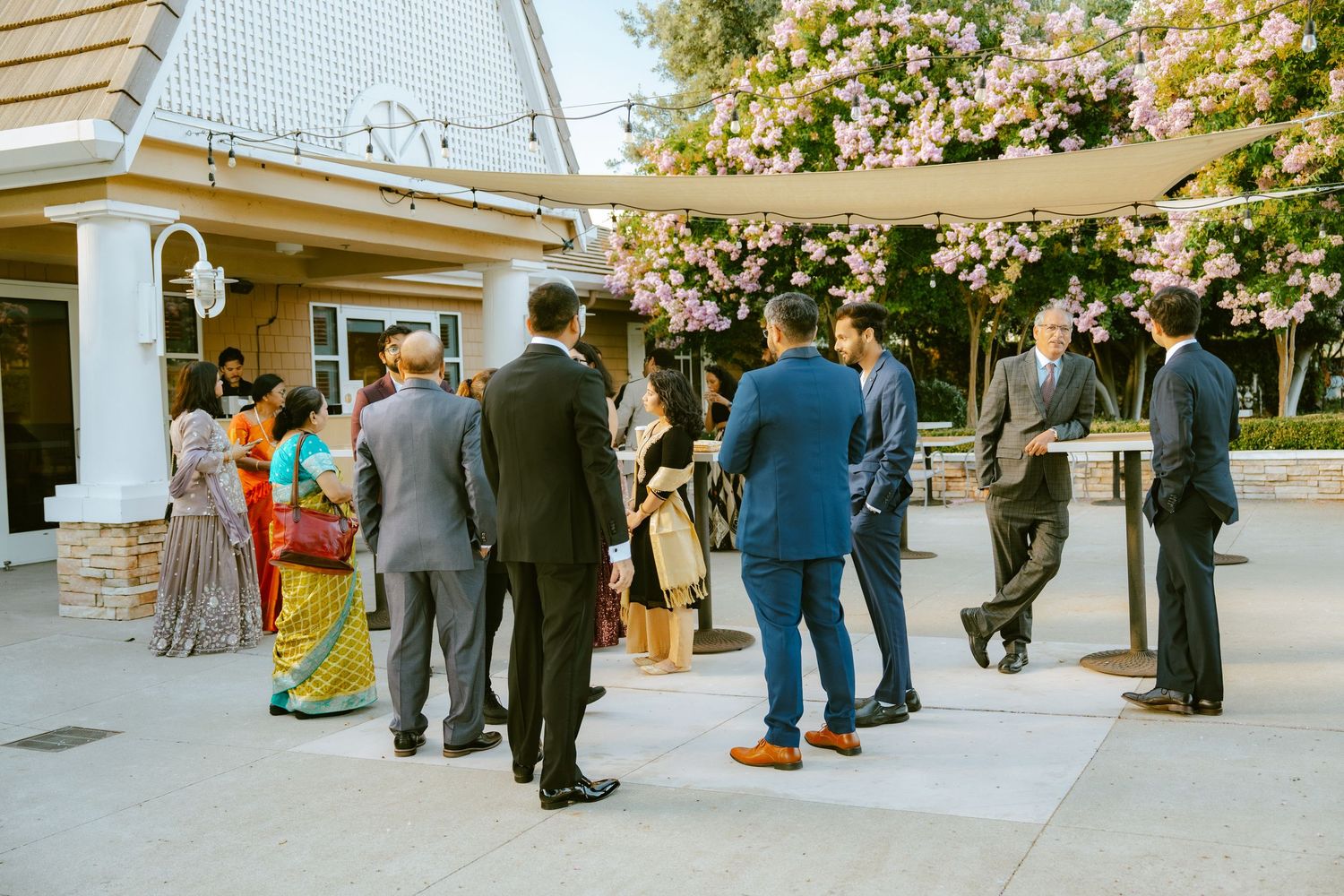 Guests in formal attire mingle outdoors at a wedding reception near a blooming tree and elegant venue.