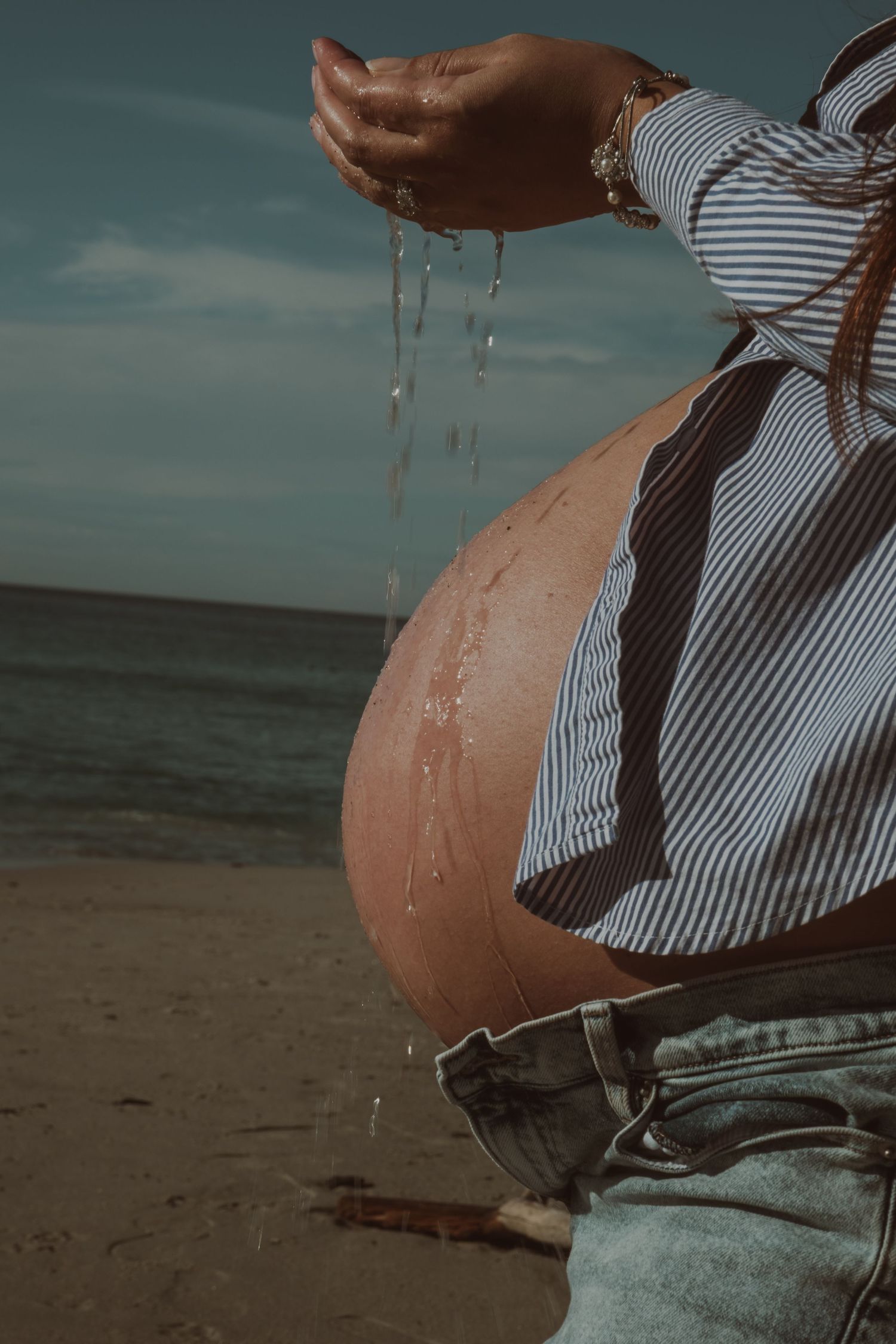 Close-up of a pregnant belly in striped clothing against a beach backdrop.