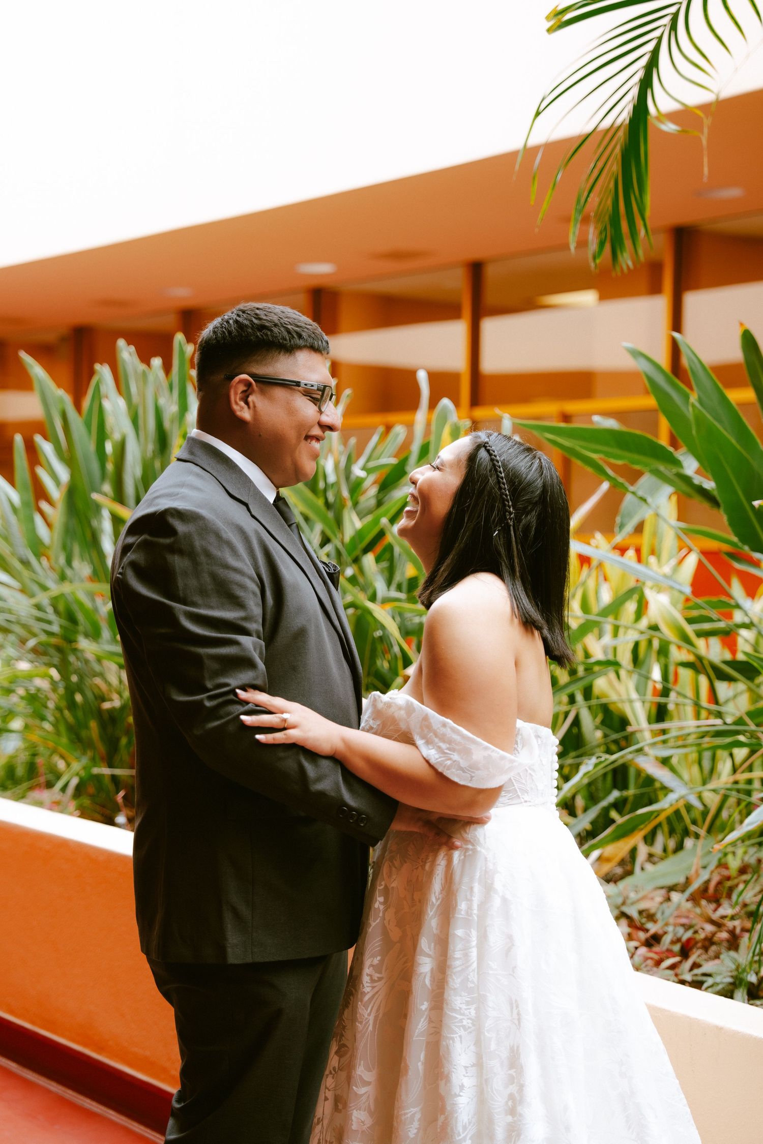 A bride in a white off-shoulder gown laughs joyfully with her groom in a dark suit, surrounded by tropical greenery.